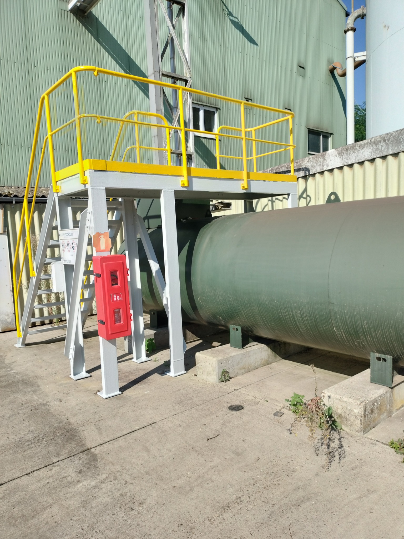 Metalowa konstrukcja schodów z żółtymi barierkami prowadząca do platformy nad zielonym zbiornikiem przemysłowym, z czerwoną szafką przeciwpożarową na podeście, na tle blaszanej ściany budynku.