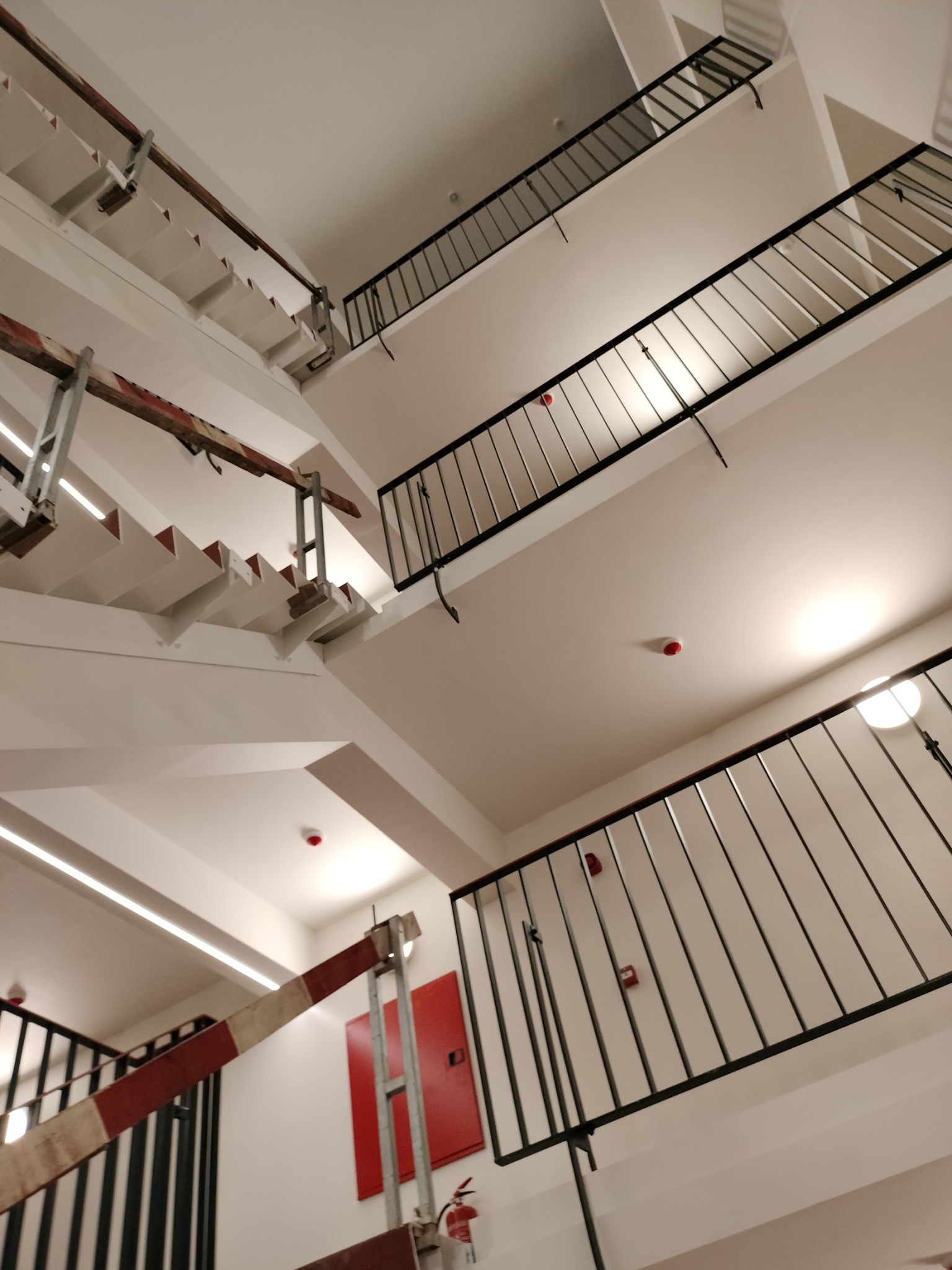 Klatka schodowa w budynku, widok z dołu, białe ściany i schody, czarne metalowe balustrady, widoczne zabezpieczenia budowlane z czerwono-białych pasów.