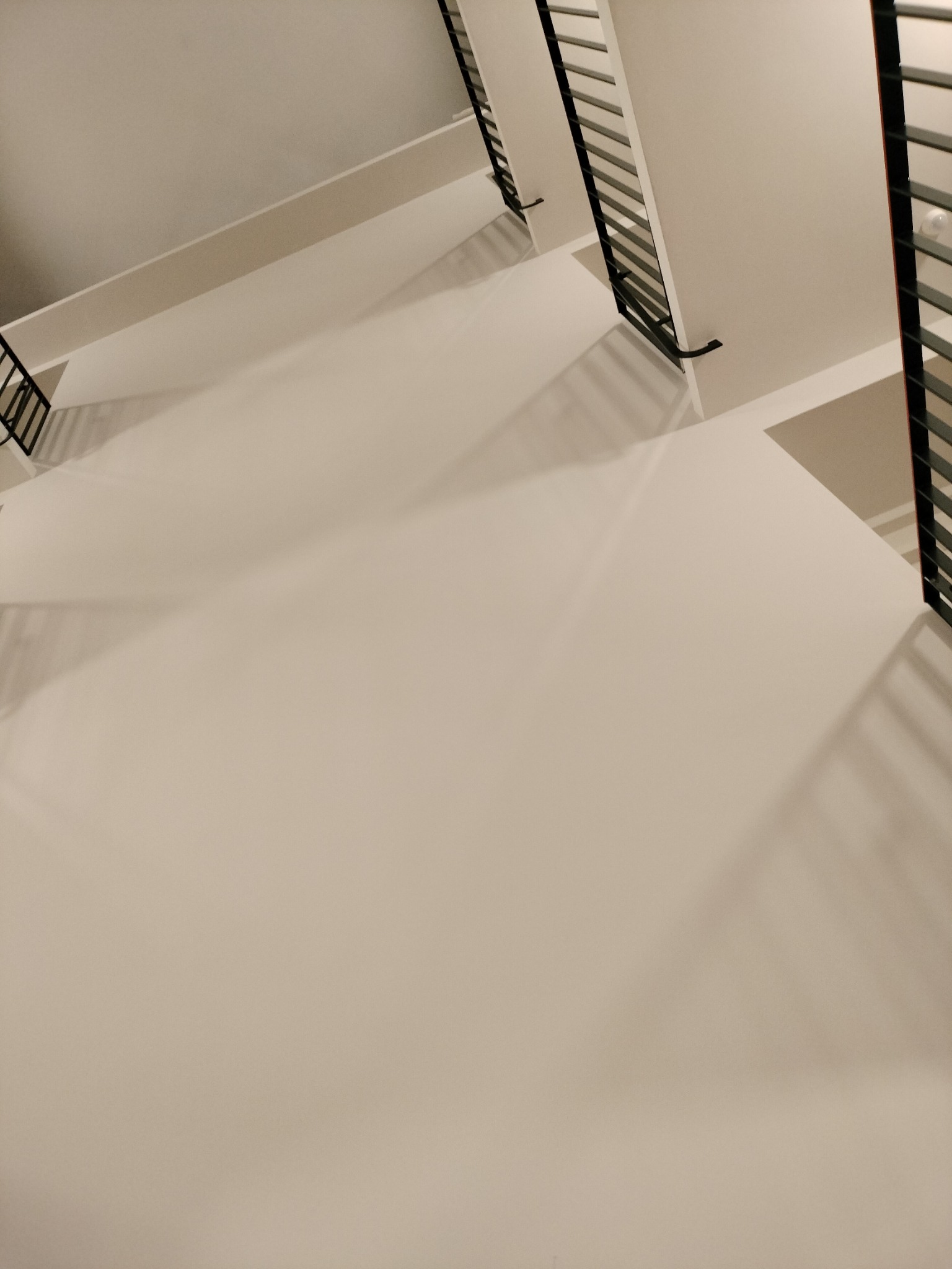 Widok z góry na klatkę schodową z białymi ścianami i czarnymi metalowymi balustradami, oświetloną ciepłym światłem.