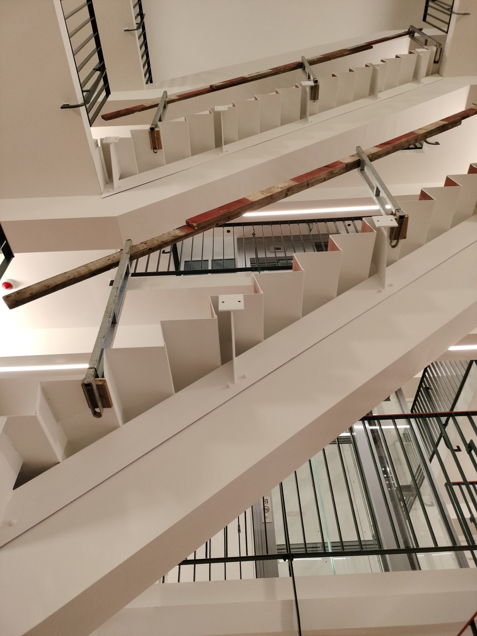 Białe, nowoczesne schody z metalowymi balustradami i tymczasowym zabezpieczeniem z drewnianych belek z czerwonymi oznaczeniami, widok z dołu na konstrukcję.