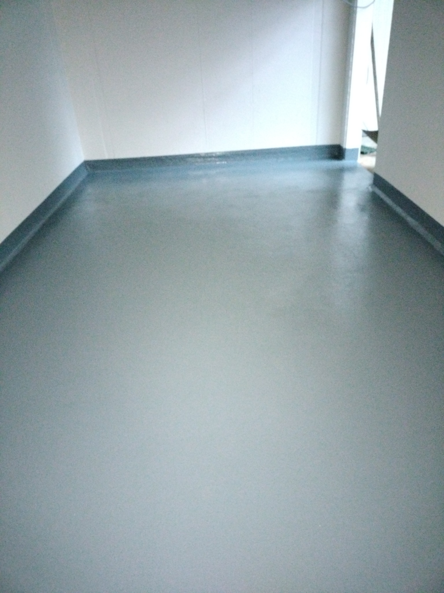 Świeżo wylana, szara, samopoziomująca wylewka podłogowa w pomieszczeniu z białymi ścianami i szarymi listwami przypodłogowymi, widoczny fragment otwartych drzwi w tle.