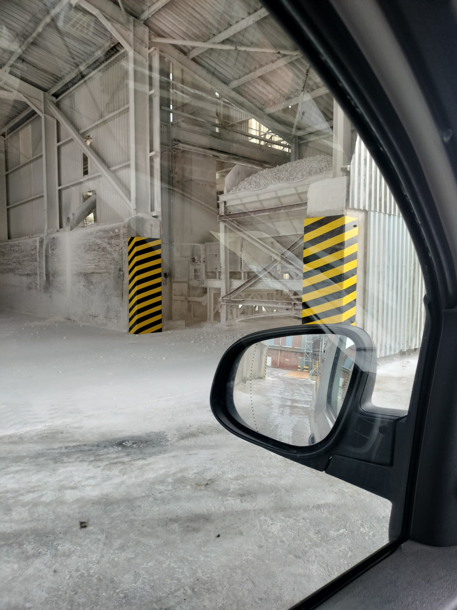 Widok z samochodu na wnętrze hali produkcyjnej z konstrukcją stalową, betonowymi elementami i hałdą białego materiału, prawdopodobnie gipsu, z kolumnami w czarno-żółte pasy.