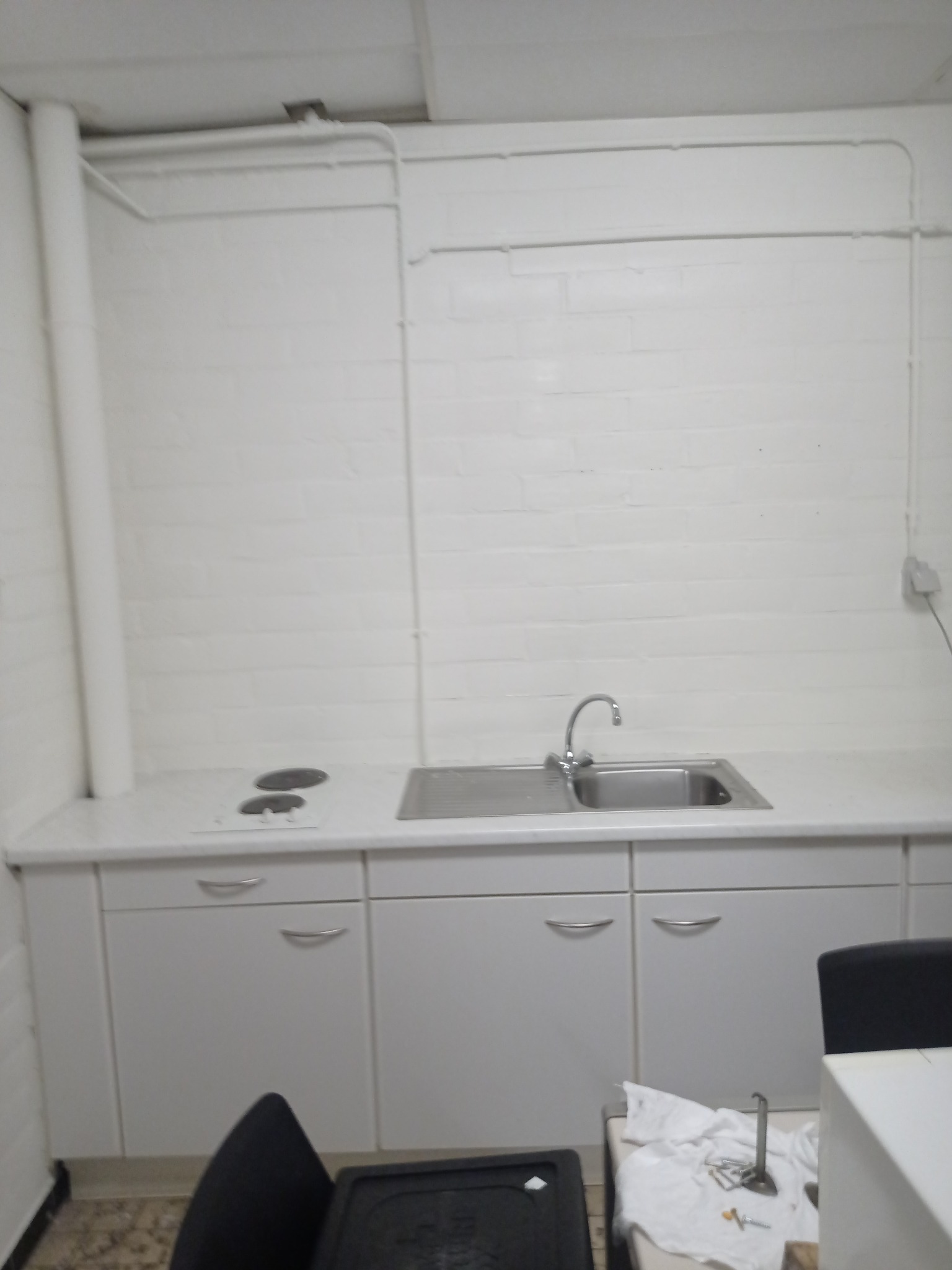Biała kuchnia z umywalką i płytą grzewczą, widoczne rury na ścianie i narzędzia na blacie, prawdopodobnie w trakcie remontu.
