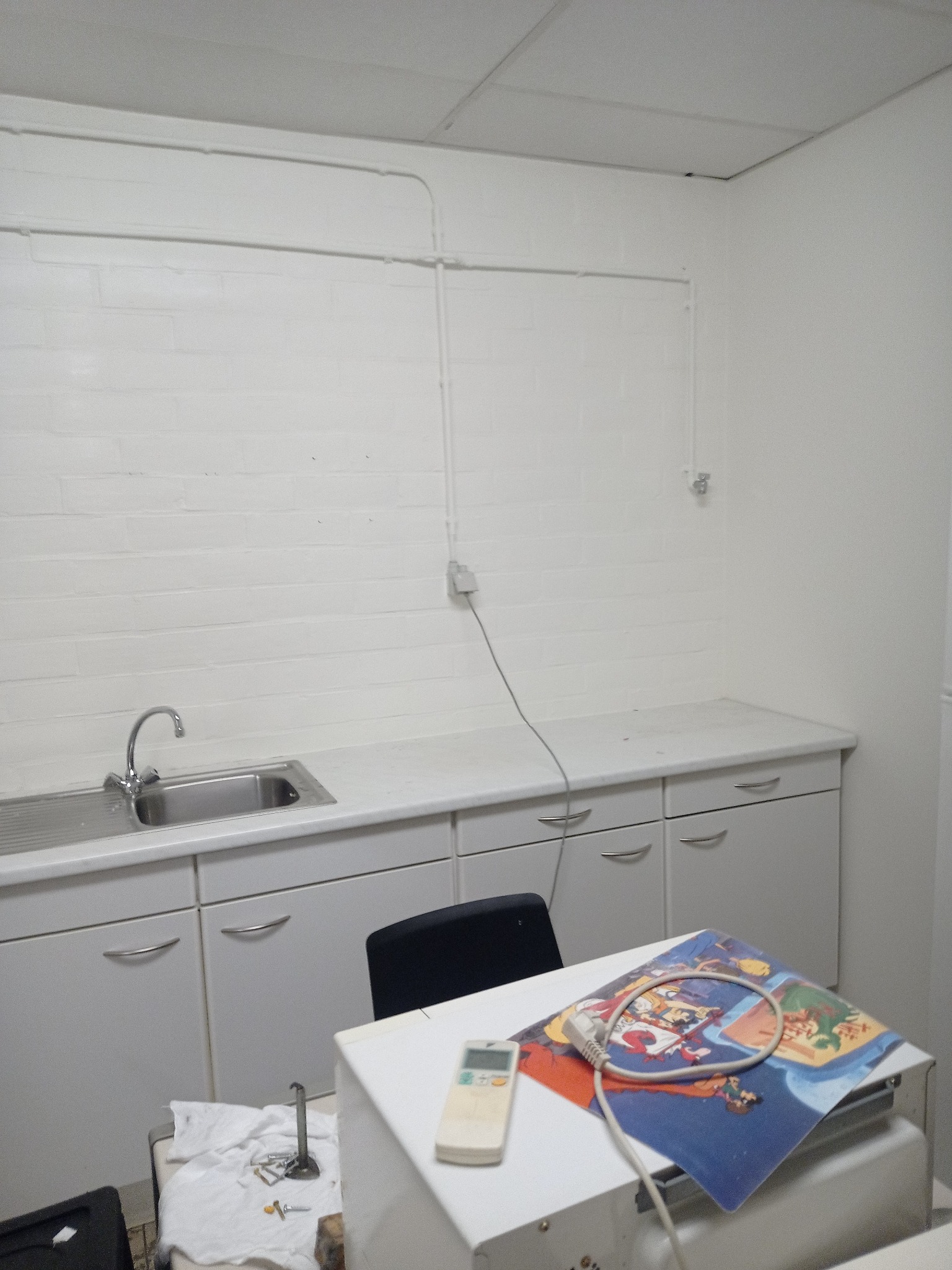 Wnętrze pomieszczenia z białą zabudową kuchenną, zlewem, kranem i widocznymi rurami na ścianie, na blacie leży kolorowa podkładka pod mysz z motywem bajkowym i pilot do klimatyzacji.