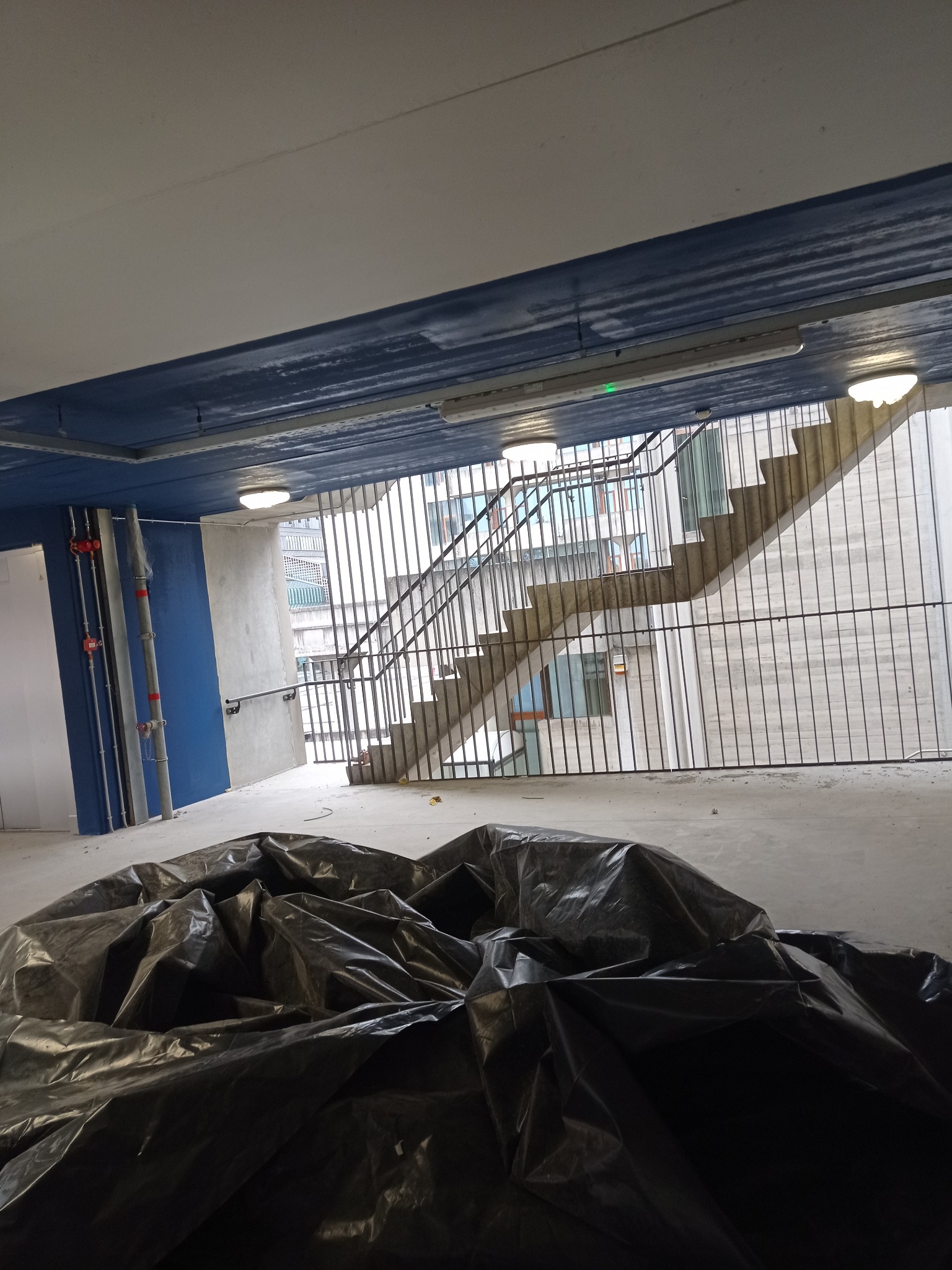 Wnętrze w trakcie remontu: betonowe schody z metalową barierką, zabezpieczone folią malarską, ściany pomalowane na niebiesko w górnej części, widoczne rury instalacyjne.