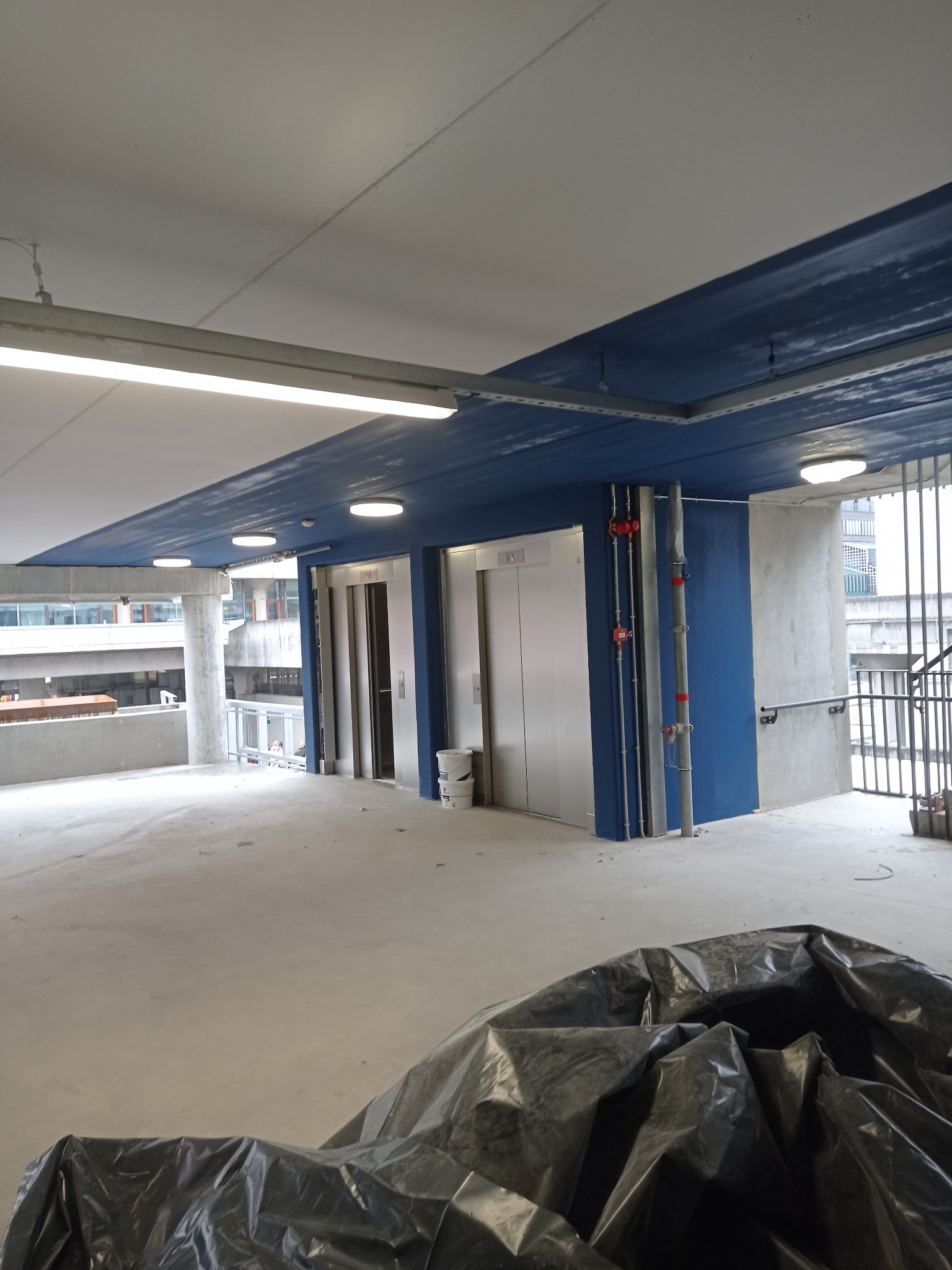 Wnętrze parkingu wielopoziomowego w trakcie prac wykończeniowych, widoczne windy, pomalowany na niebiesko sufit, betonowa podłoga, zabezpieczona folią budowlaną, oraz ślady po pracach malarskich.