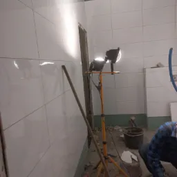 Układanie białych, błyszczących płytek w łazience, widoczna lampa budowlana na tr&oacute;jnogu, wiadro z narzędziami i fragment osoby w niebieskiej koszuli w kratę.