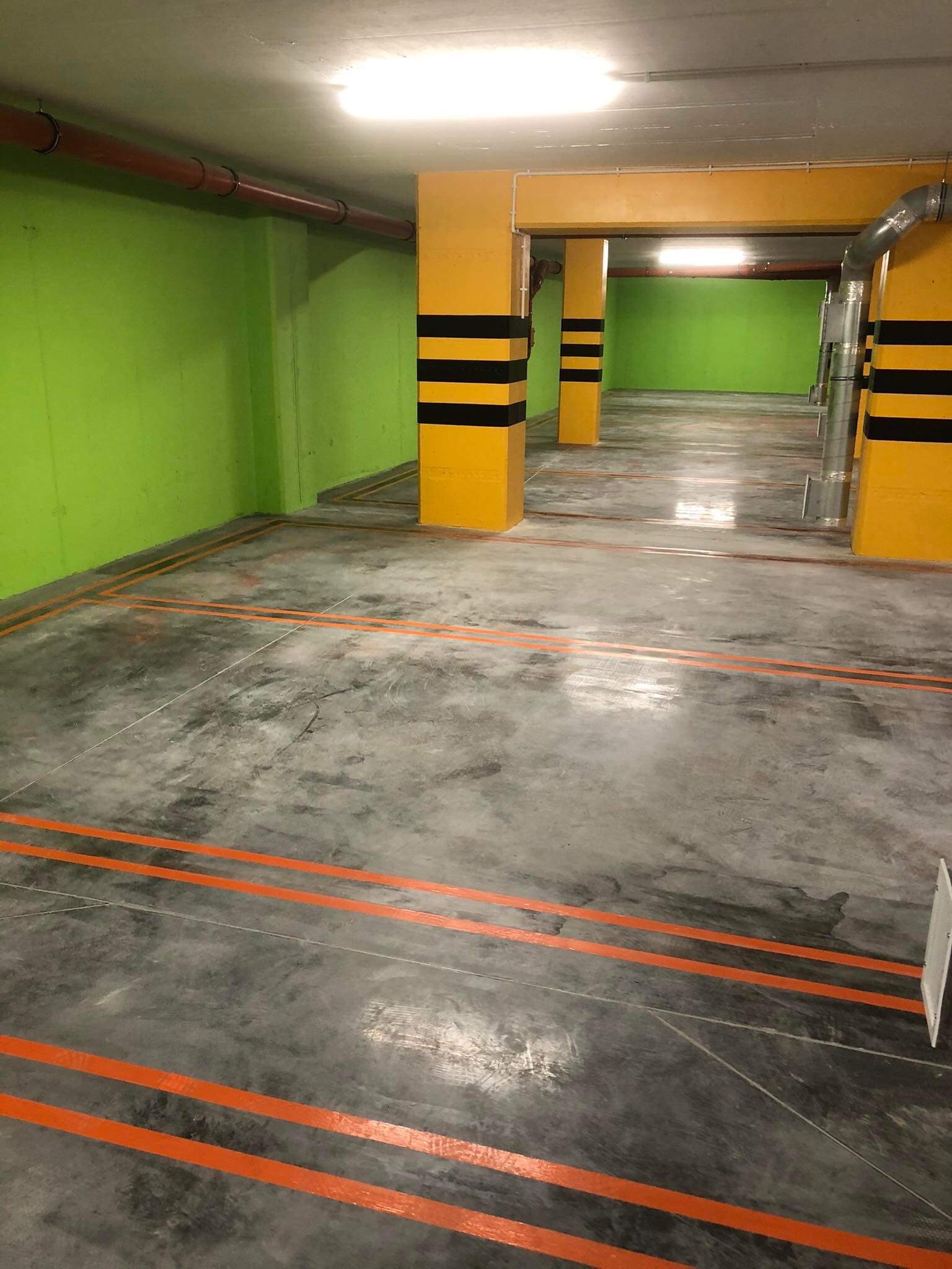 Oznakowane pomarańczowymi liniami, szare betonowe podłoże w garażu podziemnym z zielonymi ścianami i żółtymi słupami w czarne pasy.