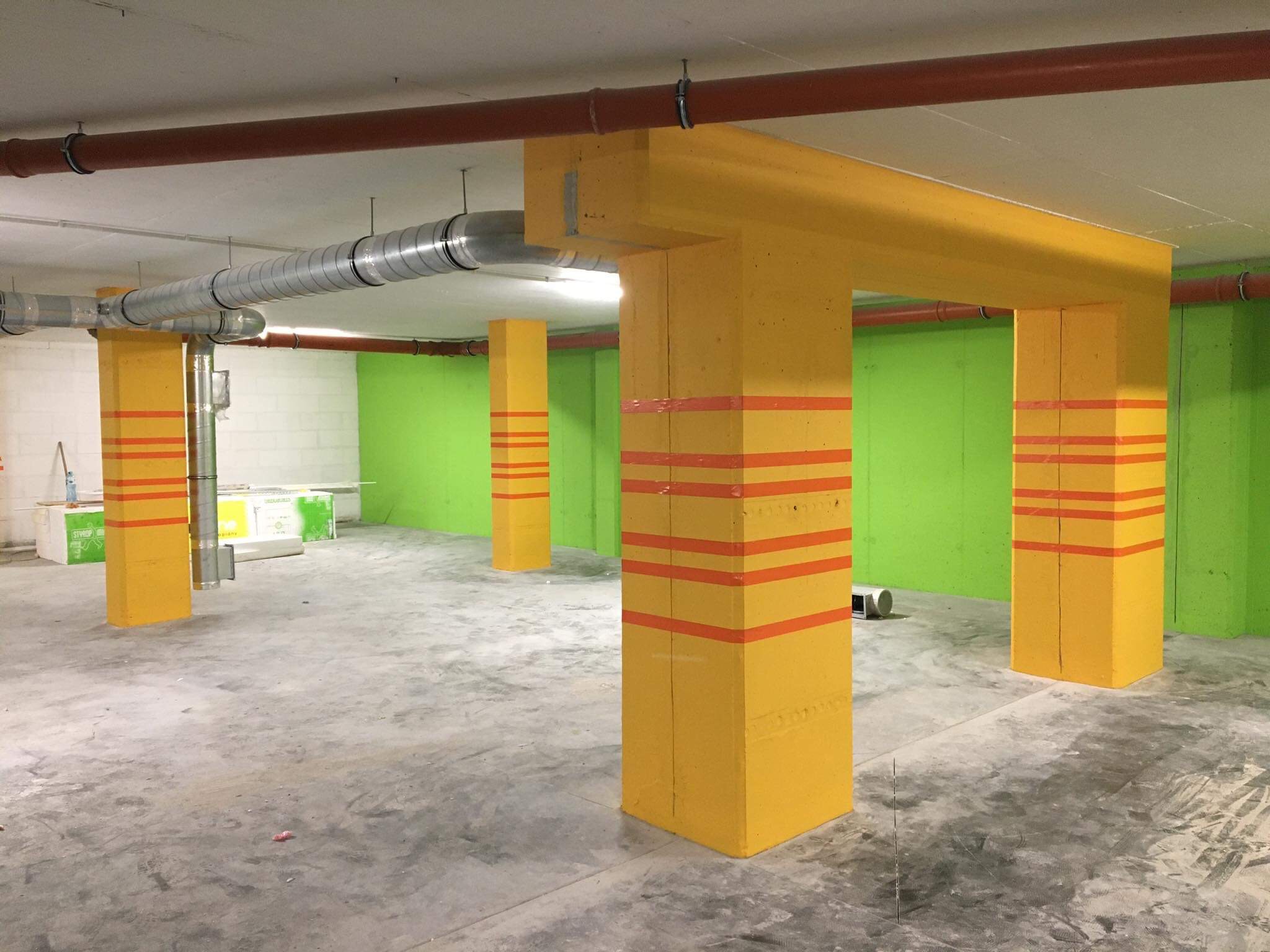 Żółte, pomalowane filary z pomarańczowymi pasami w garażu podziemnym z zielonymi ścianami, widoczne rury wentylacyjne i styropian na podłodze.