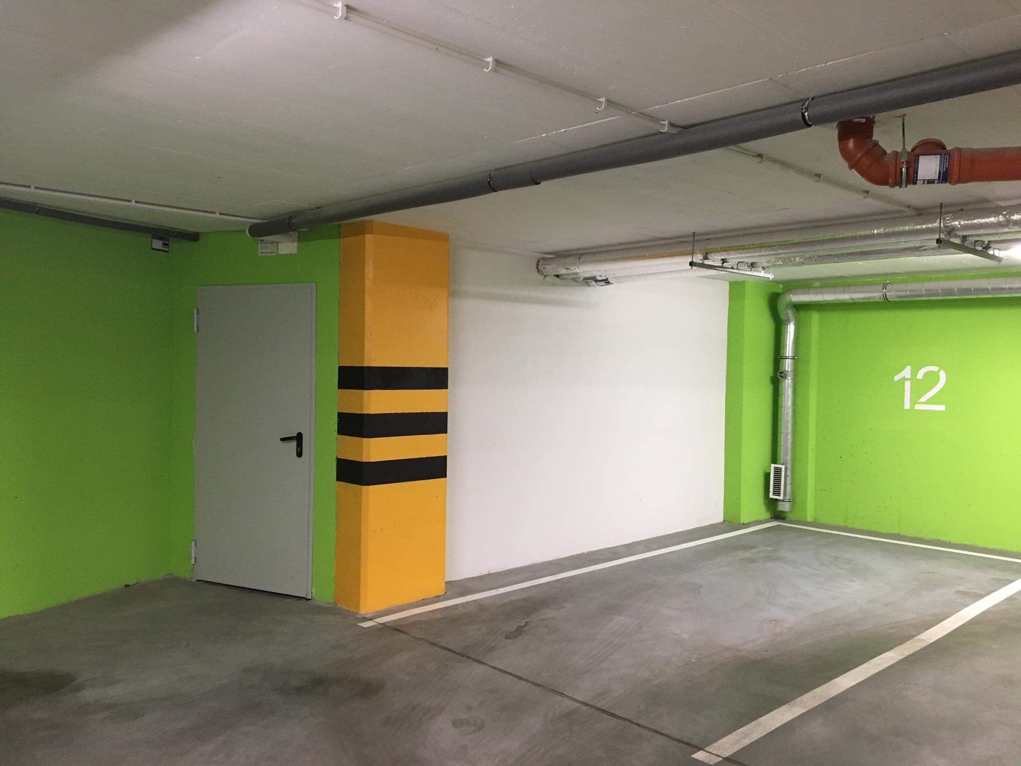 Wnętrze garażu podziemnego z pomalowanymi na zielono ścianami, żółto-czarnym słupem i szarą podłogą z wyznaczonym białym pasem miejsca parkingowego.