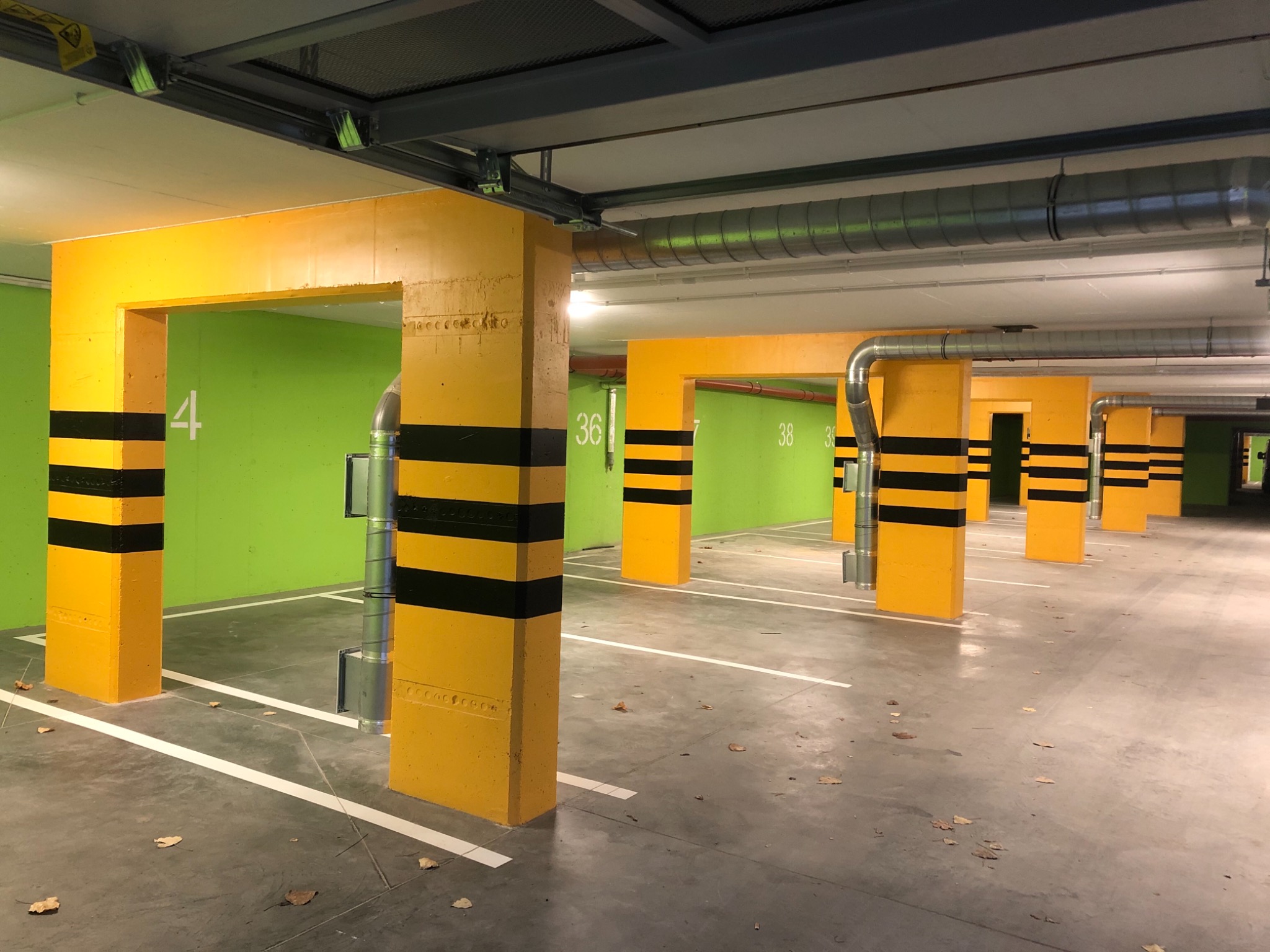 Wnętrze parkingu podziemnego z żółto-czarnymi filarami i jasnozielonymi ścianami, widoczne numery miejsc parkingowych, białe linie wyznaczające miejsca postojowe oraz metalowe rury wentylacyjne...