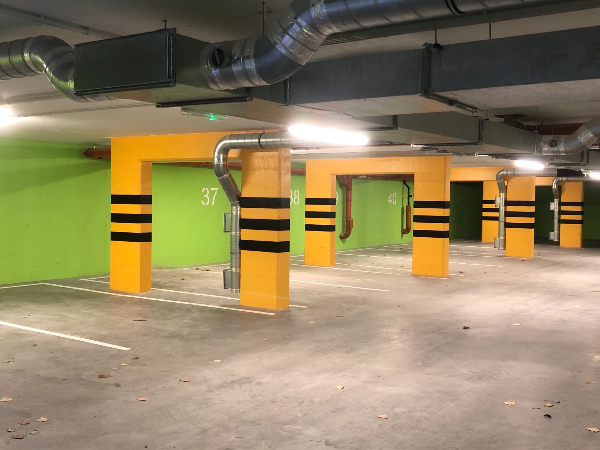 Jasnozielona ściana i żółte słupy z czarnymi pasami w garażu podziemnym, widoczne rury wentylacyjne pod sufitem.