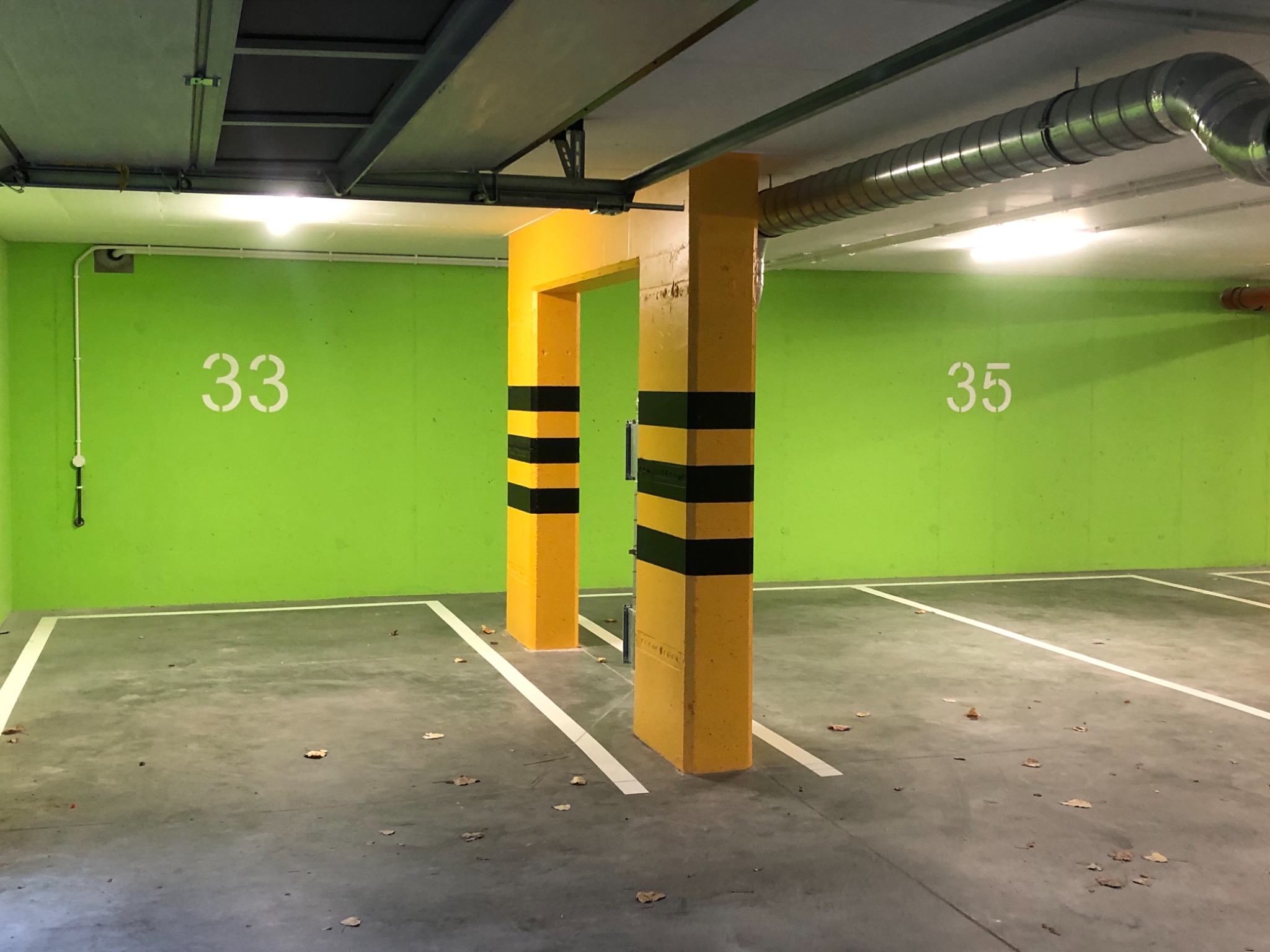 Wnętrze parkingu podziemnego w Piotrkowie Trybunalskim z jasnozielonymi ścianami oznaczonymi numerami miejsc, żółtymi słupami z czarnymi pasami i betonową podłogą pokrytą liśćmi.