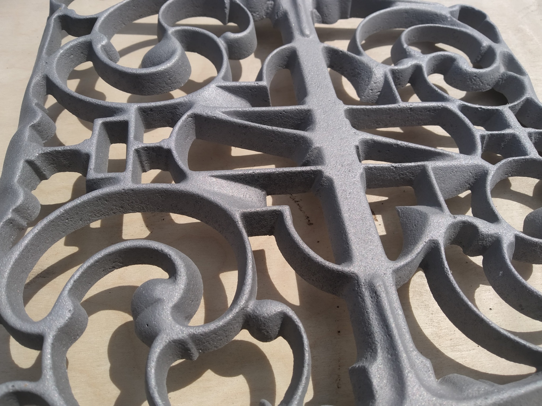 Szary, metalowy element dekoracyjny z ażurowym wzorem krzyża i spiral na jasnym, drewnianym tle, oświetlony bocznym światłem.