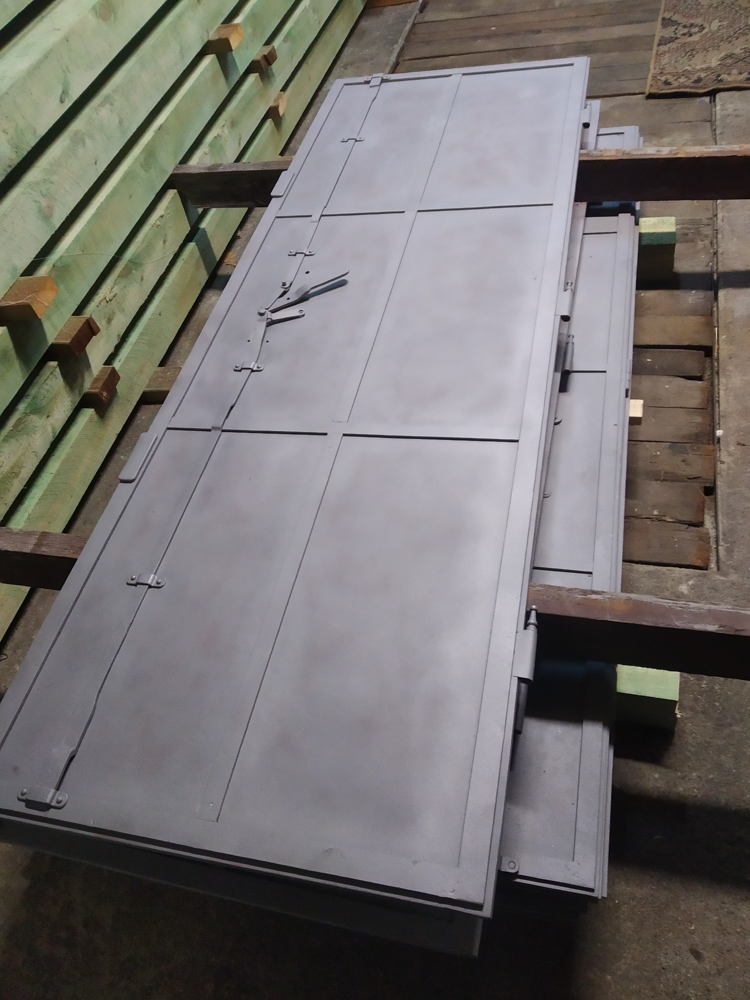 Stos szarych, metalowych drzwi garażowych z widocznymi zawiasami i zamknięciem, leżących na drewnianych belkach, po obróbce.