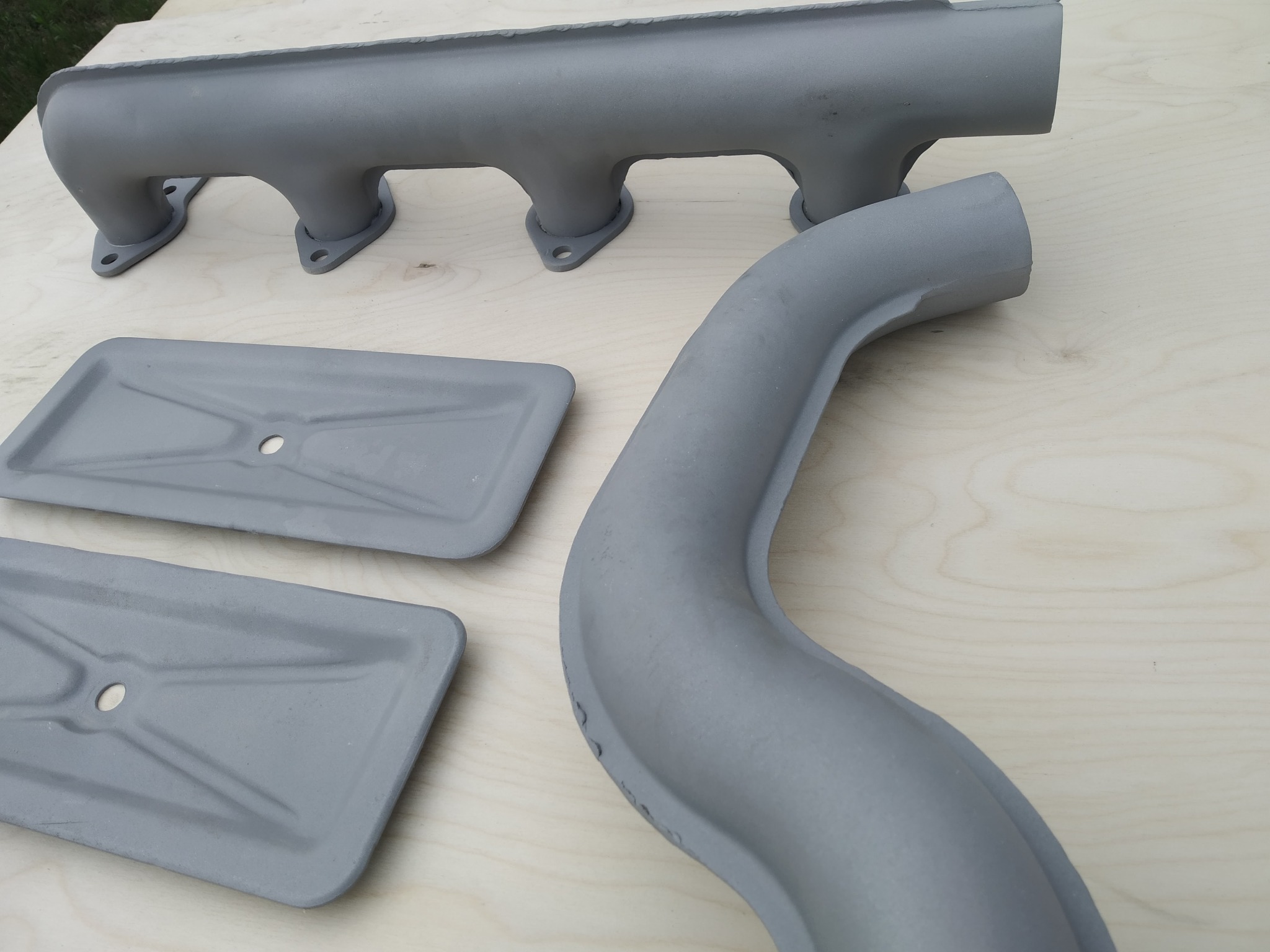 Szare, metalowe elementy samochodowe po obróbce, leżące na drewnianej powierzchni. Widoczny kolektor wydechowy, rura i dwie prostokątne płyty z otworami.