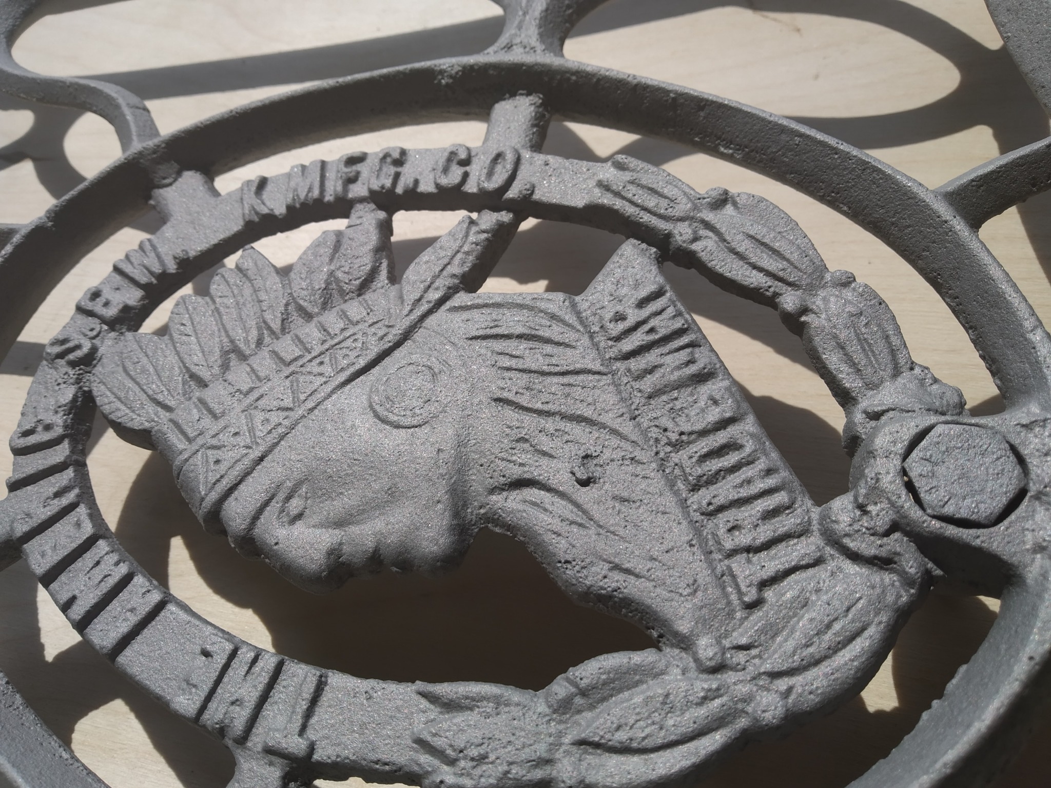 Metalowy element dekoracyjny w kształcie głowy Indianina z pióropuszem, otoczony okręgiem z napisem 'NEW MFG.CO.', po obróbce ściernej.