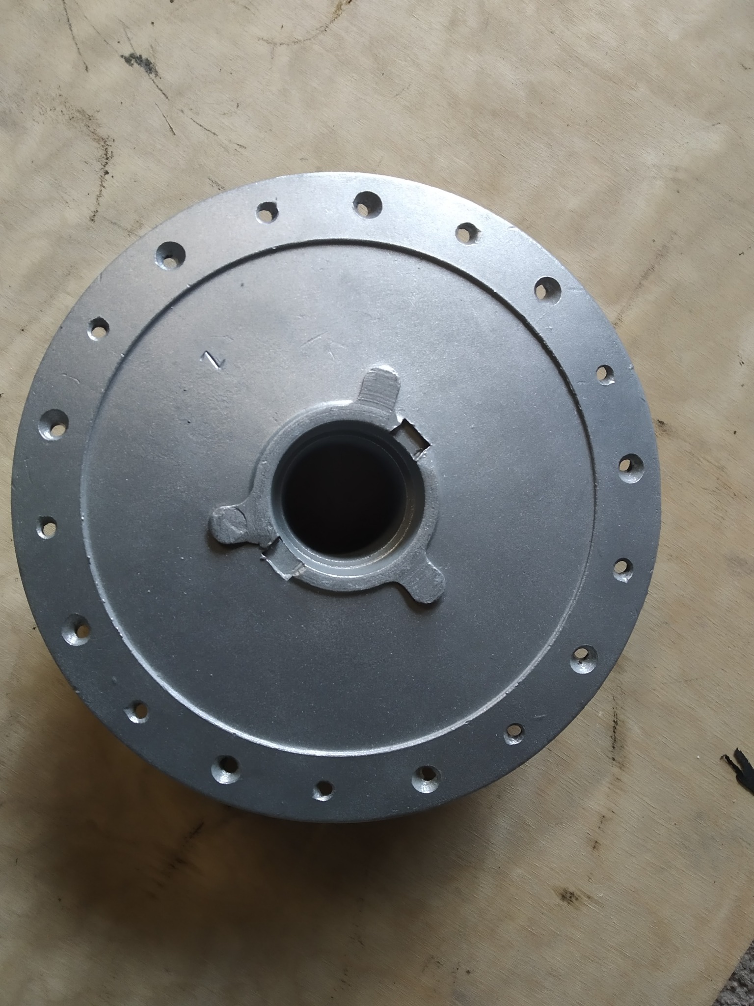 Szara, okrągła piasta koła z otworami na śruby, leżąca na drewnianej powierzchni. Widoczny centralny otwór z trzema wypustkami.