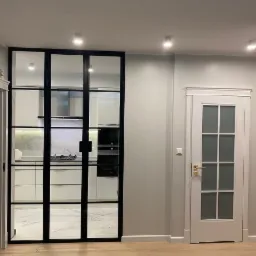 Nowoczesne, przeszklone drzwi wewnętrzne z czarnymi szprosami, oddzielające kuchnię od reszty domu. Obok widoczne białe drzwi z szybą i złotą klamką. Podłoga wyłożona panelami w odcieniach jasnego...