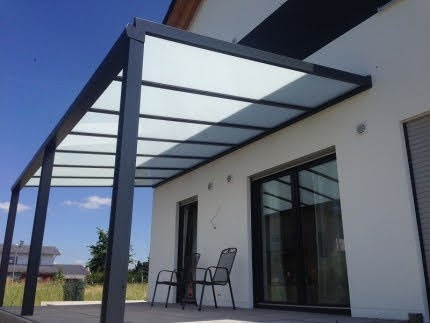 Nowoczesna pergola z szarymi słupami i mlecznym zadaszeniem przylegająca do białego budynku z dużymi oknami, uzupełniona dwoma krzesłami na zewnątrz.