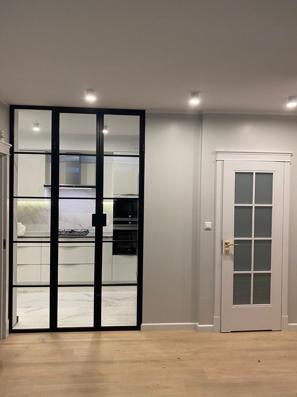 Nowoczesne, przeszklone drzwi wewnętrzne z czarnymi szprosami, oddzielające kuchnię od reszty domu. Obok widoczne białe drzwi z szybą i złotą klamką. Podłoga wyłożona panelami w odcieniach jasnego...