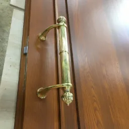 Ciemnobrązowe drewniane drzwi z ozdobną, złotą, pionową klamką z dwoma uchwytami i zdobieniami w kształcie kuli na końcach, widoczny zamek wpuszczany.