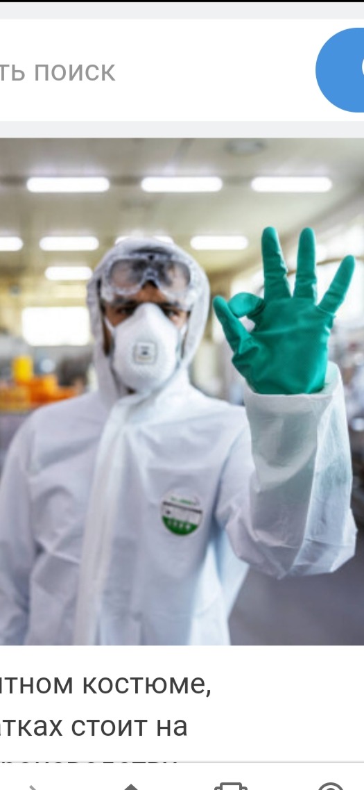 Pracownik w białym kombinezonie ochronnym, goglach i masce, z zielonymi rękawicami, pokazuje znak 'OK' na tle rozmytego wnętrza hali.