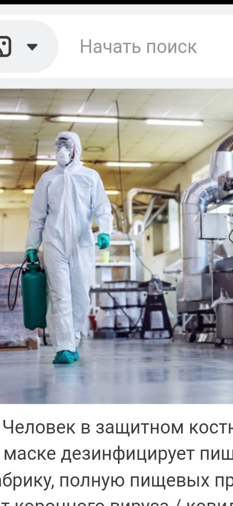 Pracownik w kombinezonie ochronnym, masce i rękawicach, niosący zielony opryskiwacz w hali produkcyjnej, prawdopodobnie podczas dezynfekcji.