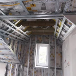 Konstrukcja metalowa pod zabudowę poddasza z widocznym ociepleniem folią paroizolacyjną Isover, drewniane belki stropowe, okno dachowe i fragment ściany z instalacją elektryczną.