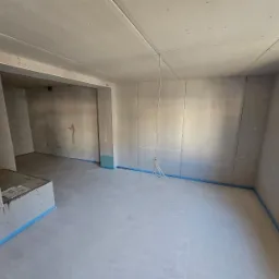 Surowy pokój z zamontowanymi płytami gipsowo-kartonowymi na ścianach i suficie, zabezpieczony niebieską taśmą malarską przy podłodze, widoczne przewody elektryczne zwisające z sufitu.