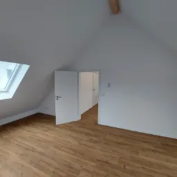 Poddasze po remoncie z drewnopodobną podłogą, białymi ścianami i otwartymi drzwiami do kolejnego pomieszczenia. W dachu okno połaciowe.