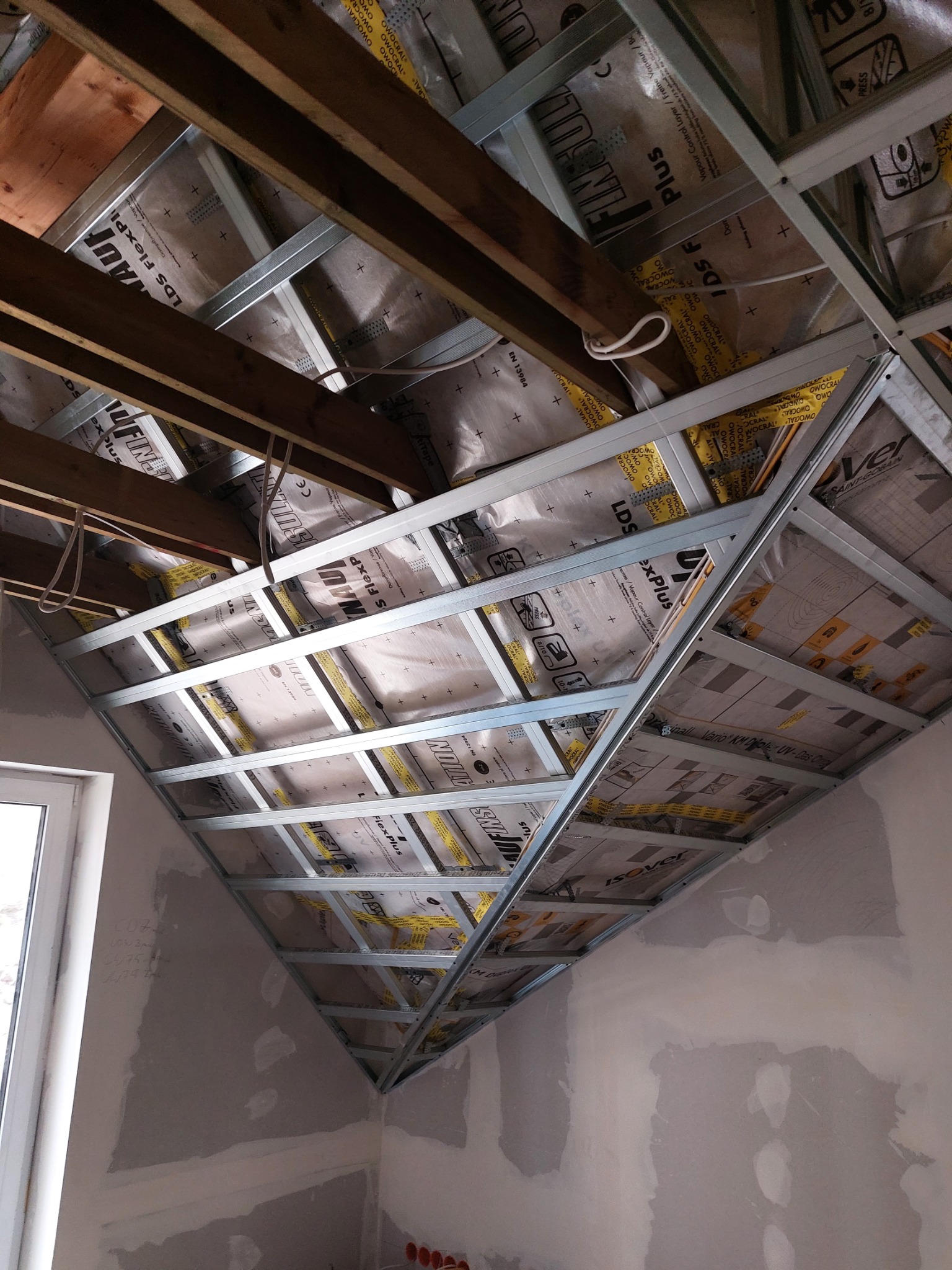 Skośne poddasze w trakcie prac izolacyjnych, widoczne drewniane belki stropowe, metalowy stelaż pod przyszłą zabudowę, folia paroizolacyjna z nadrukiem, niedokończone ściany z widocznymi śladami...