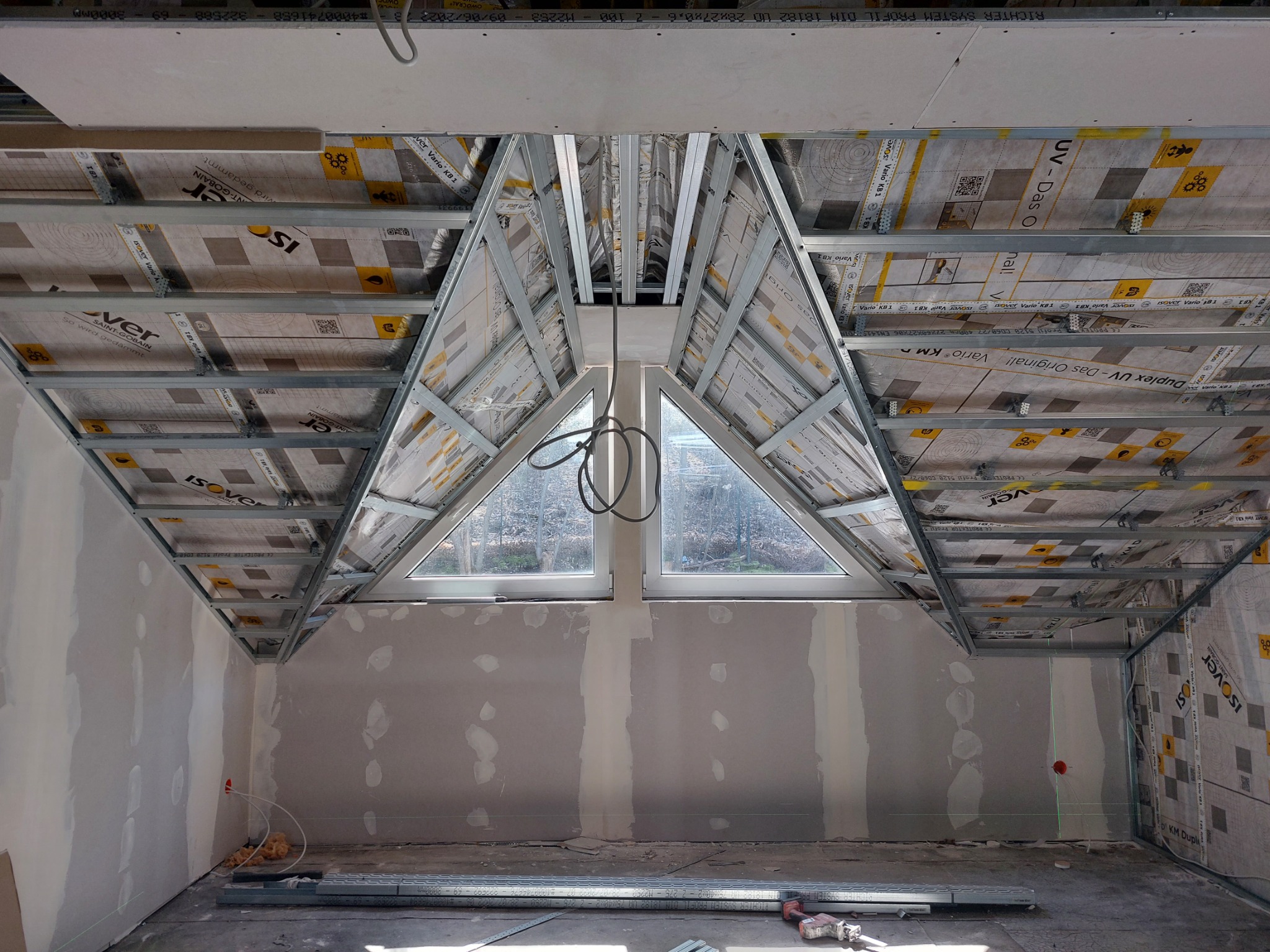 Poddasze w trakcie prac wykończeniowych: widoczna konstrukcja metalowa, izolacja termiczna Isover Vario KB1, częściowo zamontowane płyty gipsowe, okno trójkątne, ściany przygotowane do malowania...