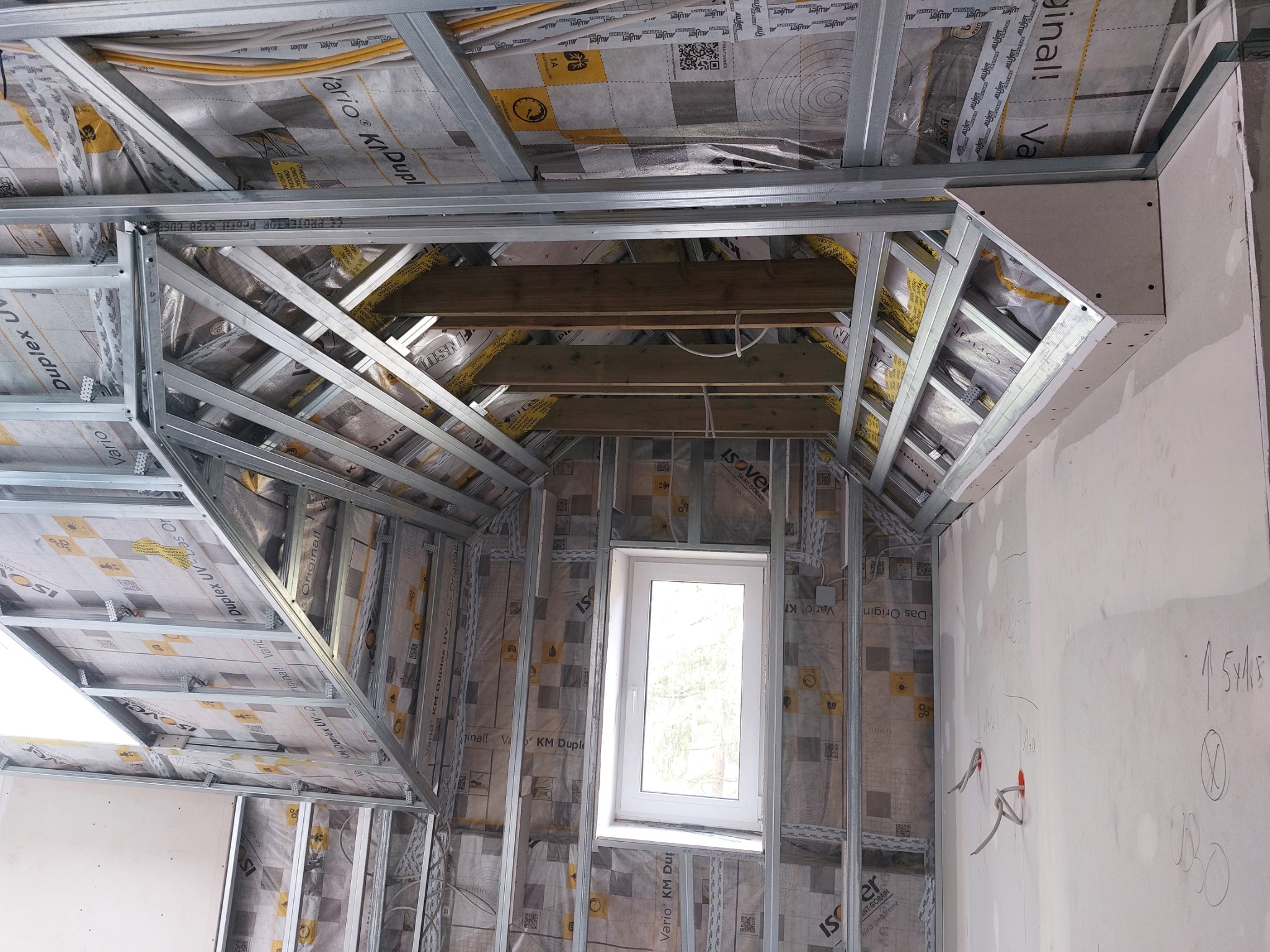 Konstrukcja metalowa pod zabudowę poddasza z widocznym ociepleniem folią paroizolacyjną Isover, drewniane belki stropowe, okno dachowe i fragment ściany z instalacją elektryczną.