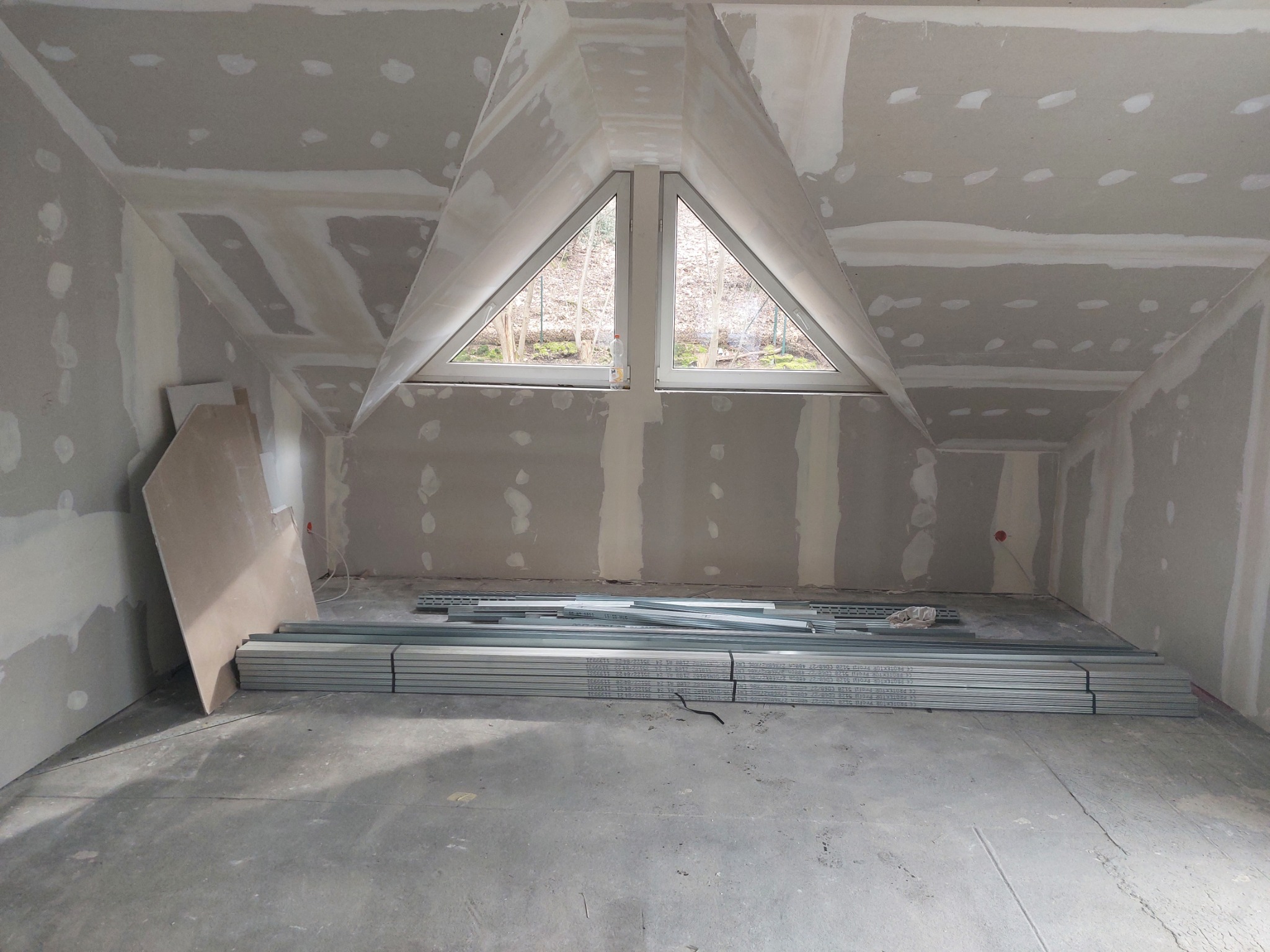 Poddasze w trakcie prac wykończeniowych: ściany i sufit pokryte płytami, widoczne szpachlowanie łączeń, profile metalowe na podłodze, trójkątne okno dachowe z widokiem na drzewa.