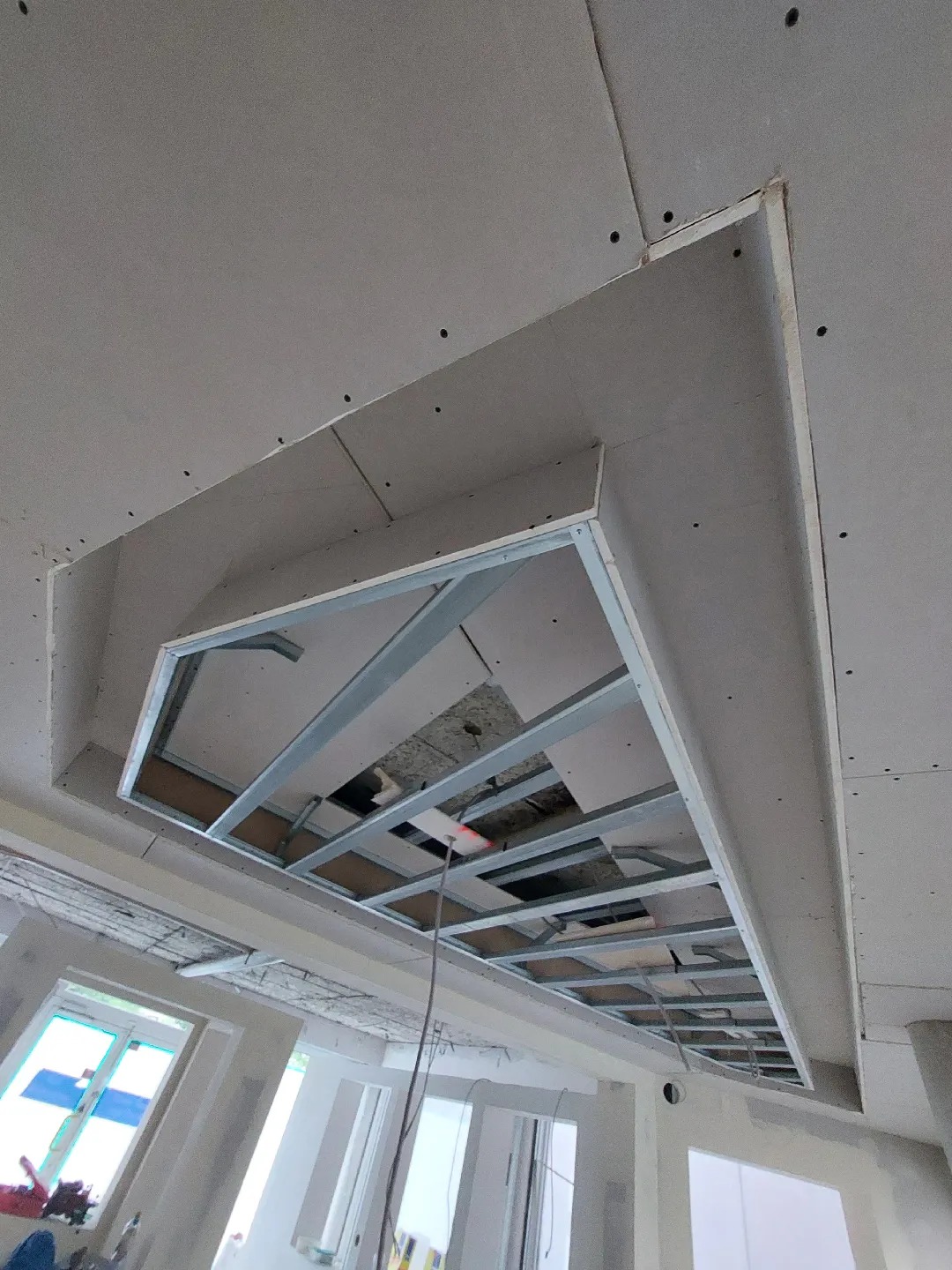 Niedokończona konstrukcja sufitu podwieszanego z metalowym stelażem i fragmentami płyt, widok z dołu, widoczne okna i fragmenty ścian w surowym stanie.