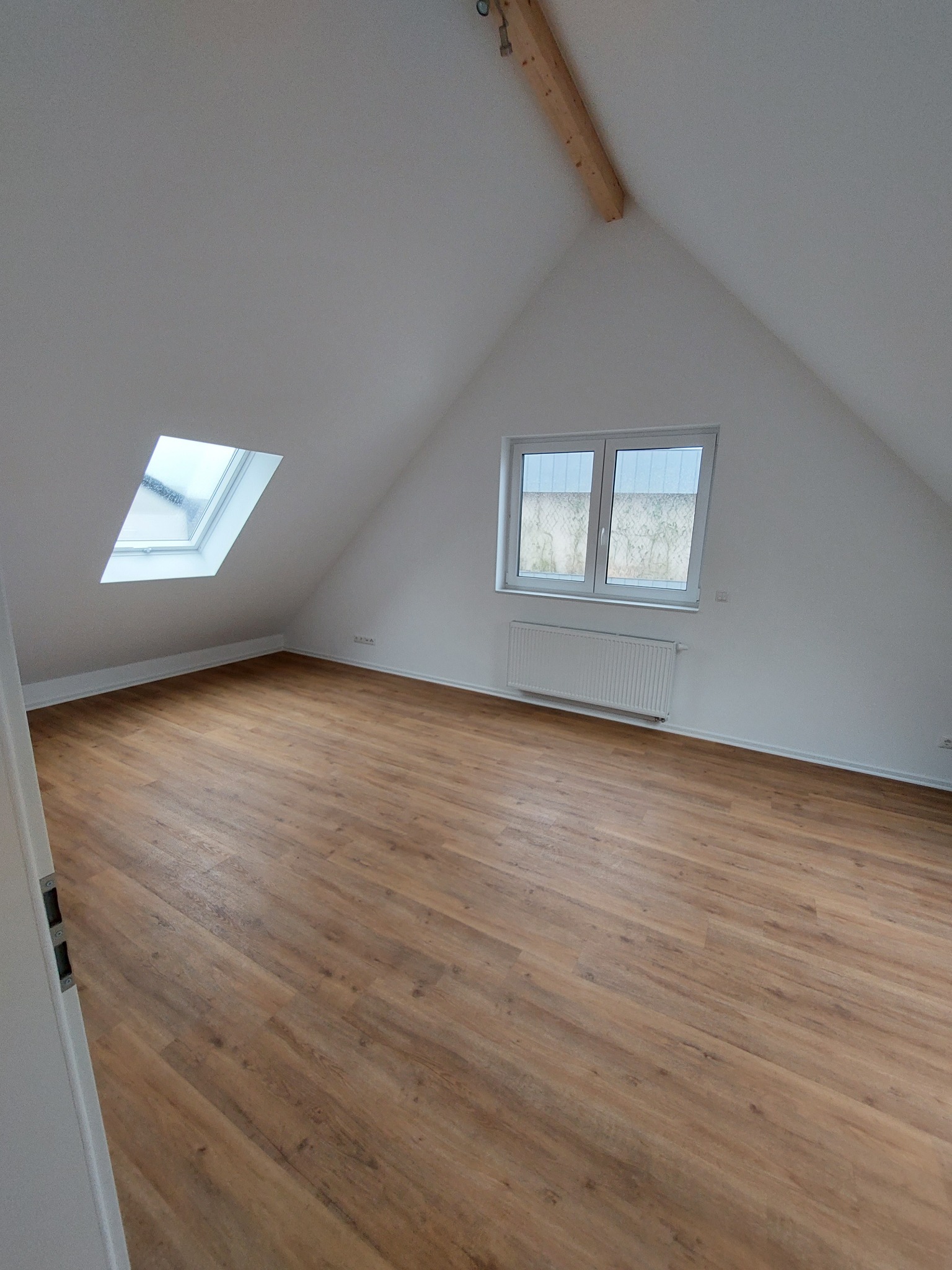 Puste poddasze ze skośnymi ścianami, drewnianą podłogą, oknem dachowym i grzejnikiem pod standardowym oknem.
