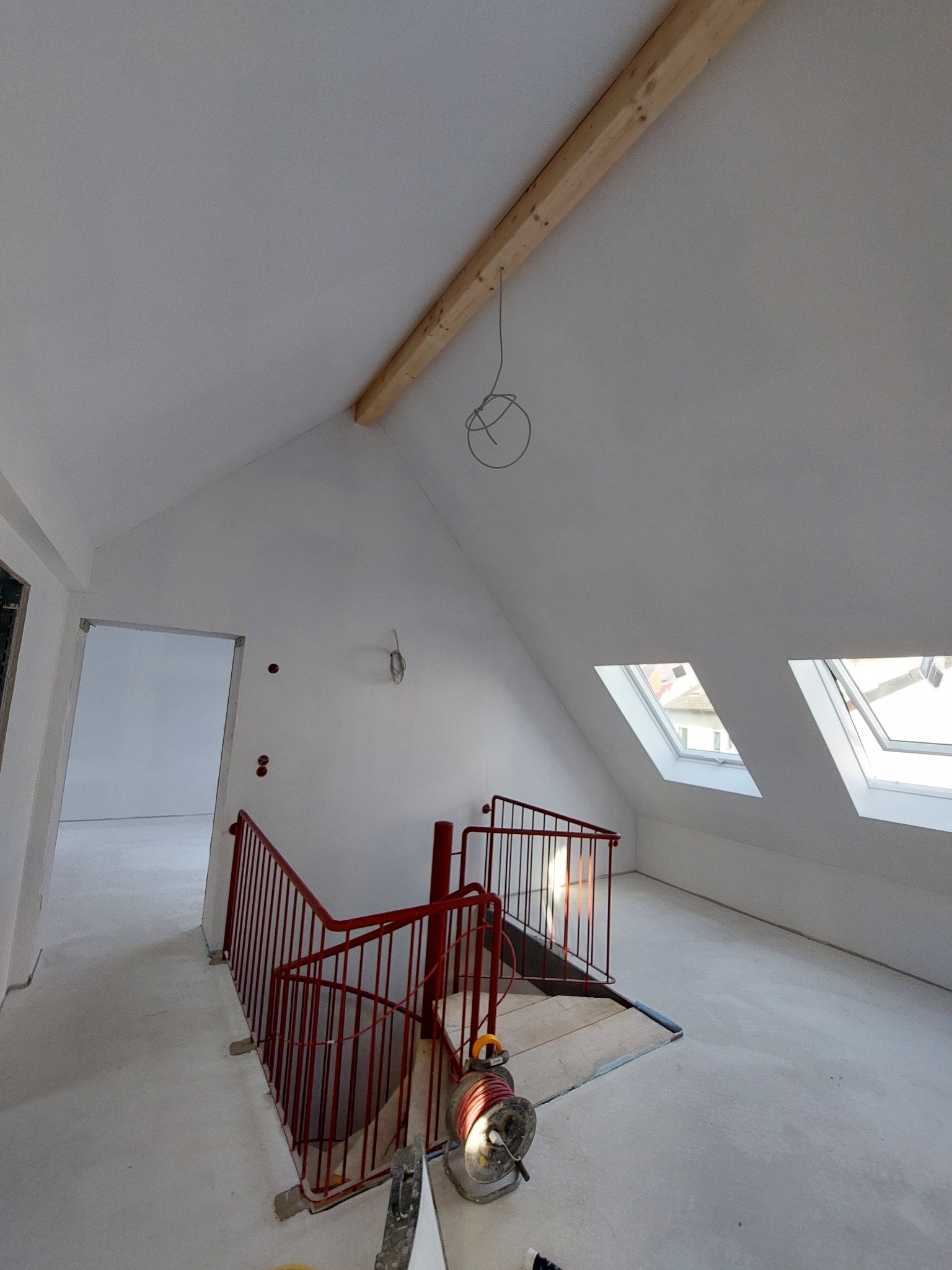 Poddasze w trakcie wykańczania: widoczne okna dachowe, czerwona balustrada schodów, surowy drewniany belka stropowa i białe ściany po szpachlowaniu, przygotowane do malowania.