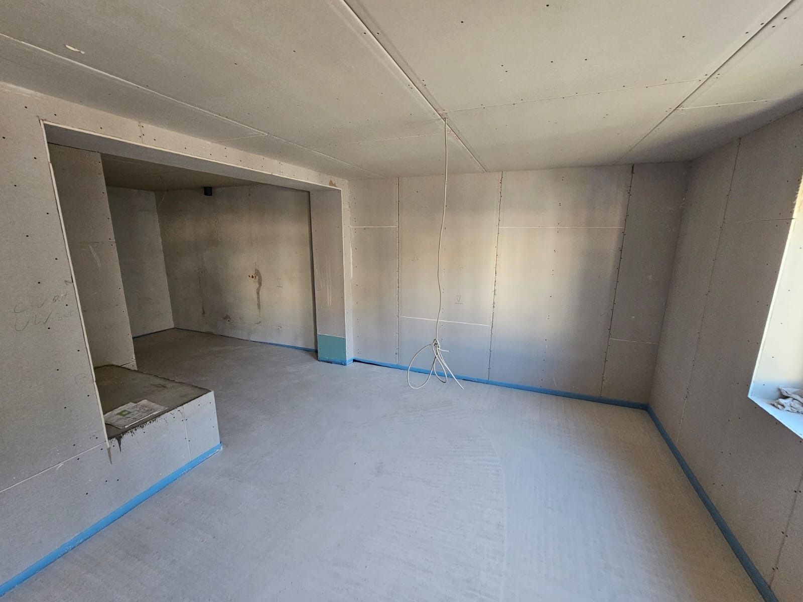 Surowy pokój z zamontowanymi płytami gipsowo-kartonowymi na ścianach i suficie, zabezpieczony niebieską taśmą malarską przy podłodze, widoczne przewody elektryczne zwisające z sufitu.