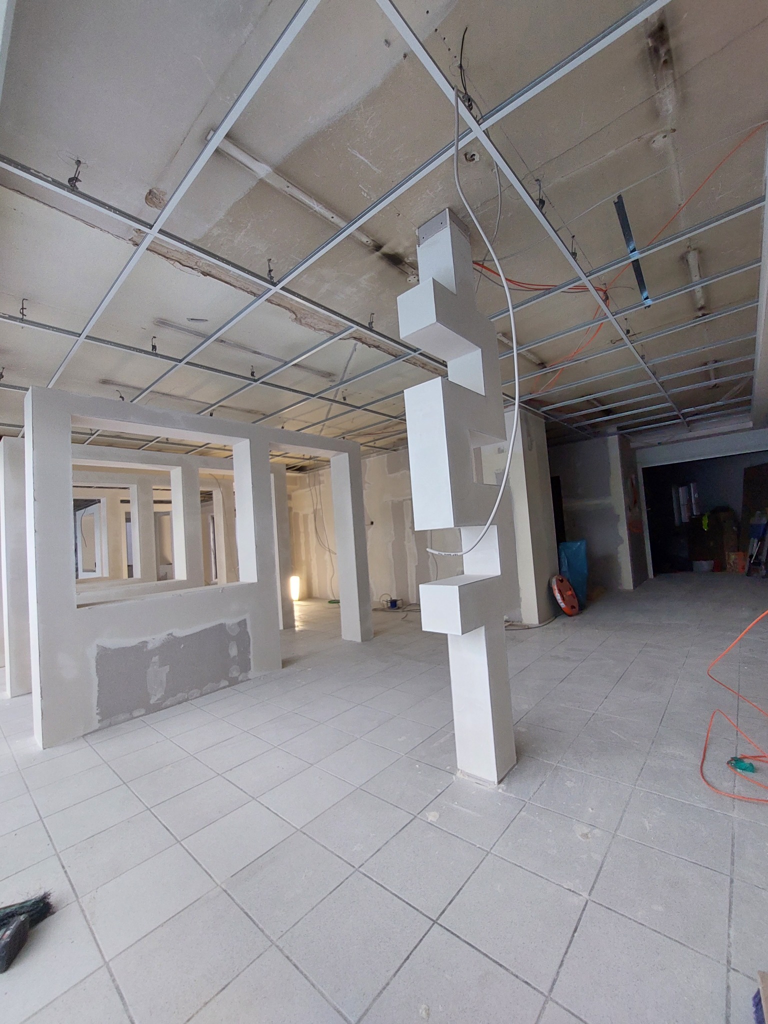 Wnętrze biurowe w trakcie prac wykończeniowych; widoczne metalowe profile sufitowe, wstępnie obrobione ściany działowe z otworami oraz dekoracyjna kolumna z białych prostopadłościanów