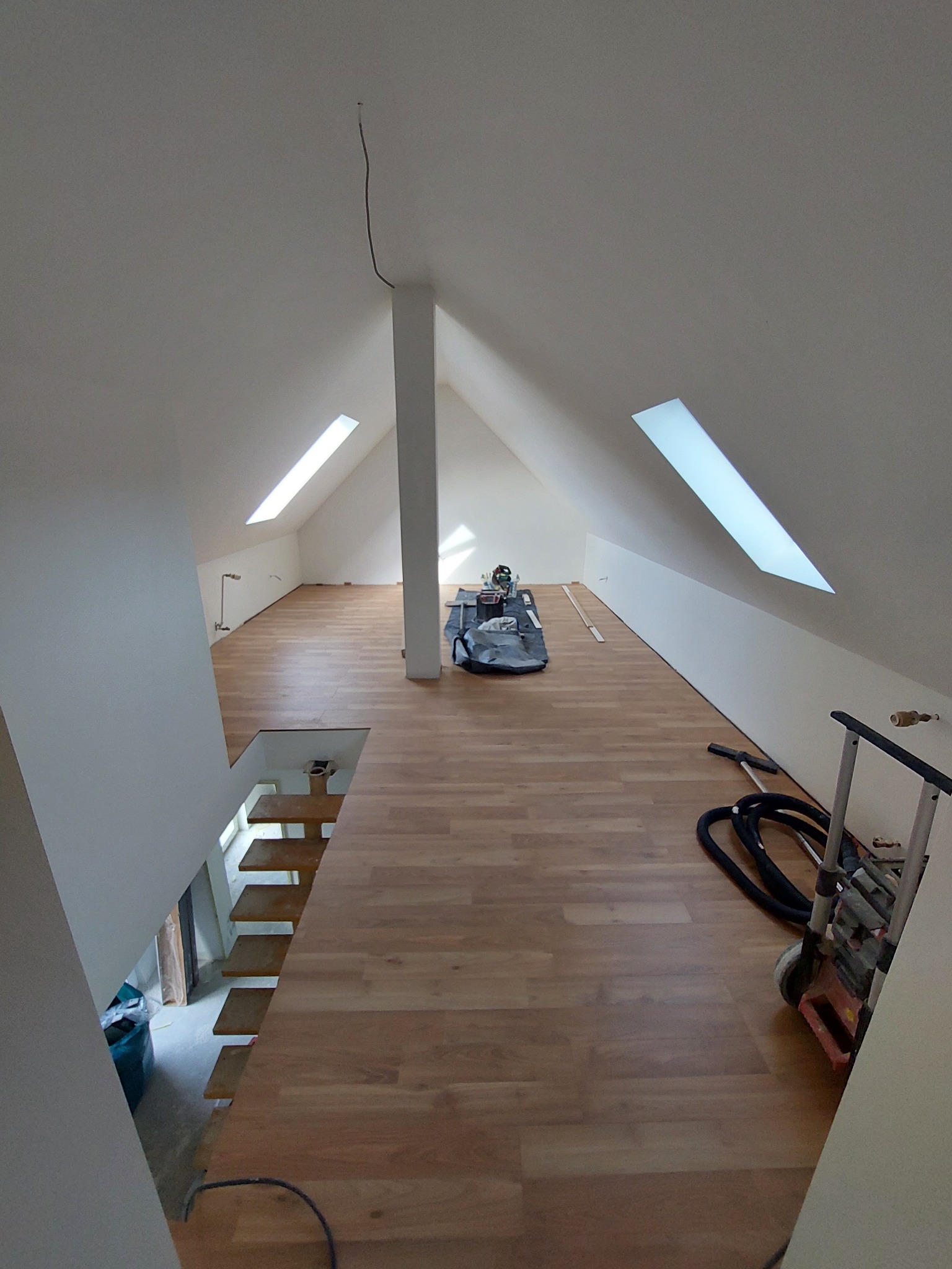 Poddasze z drewnianą podłogą w trakcie prac wykończeniowych, widoczne okna dachowe, narzędzia i maszyny budowlane, schody z drewnianymi stopniami prowadzące na niższy poziom.