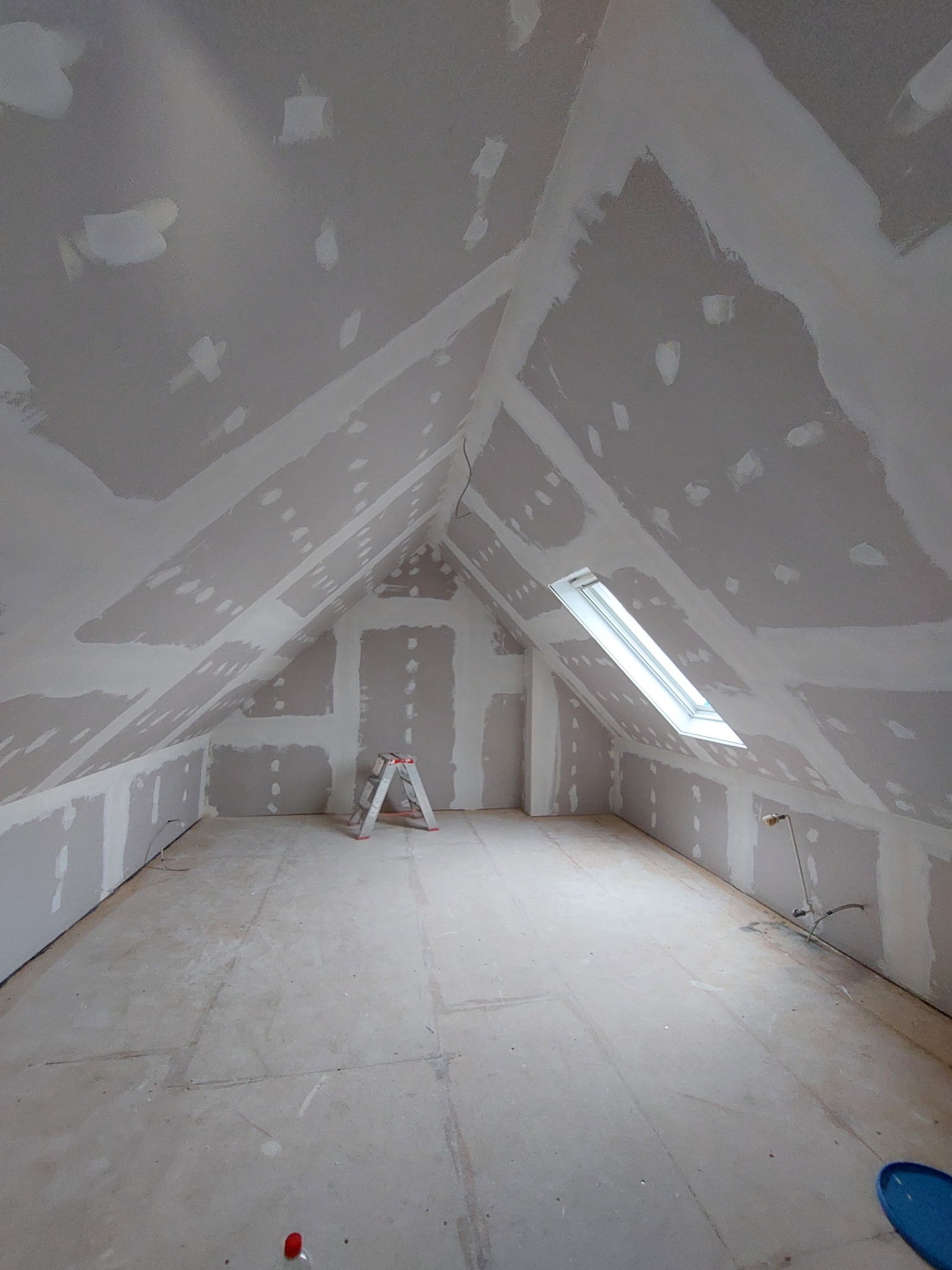 Poddasze po montażu płyt karton gips z widocznymi łączeniami i miejscami po szpachlowaniu, mała drabina rozstawiona na środku, okno dachowe w skośnej ścianie.