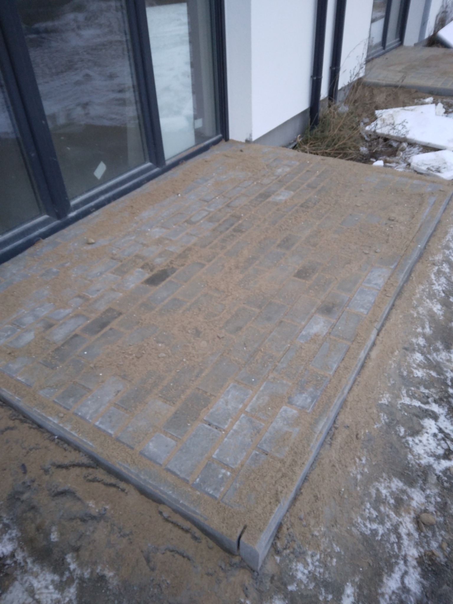 Nowo ułożona, prostokątna nawierzchnia z szarej kostki brukowej przy budynku, zasypana piaskiem, z widocznymi fragmentami śniegu.