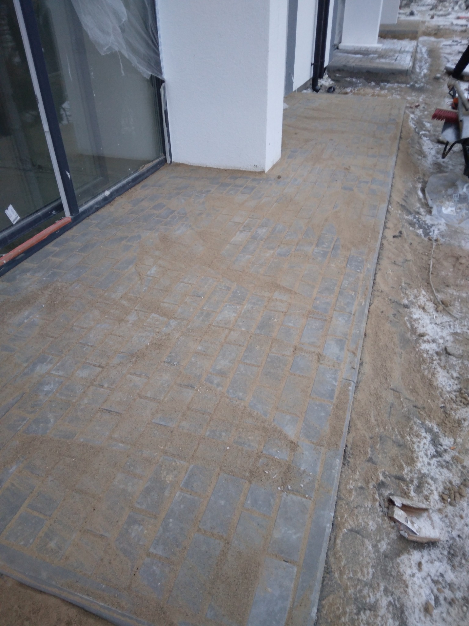 Świeżo ułożona, szara kostka brukowa przy budynku, zasypana piaskiem do wypełnienia szczelin. Widoczne fragmenty okien i elewacji budynku.