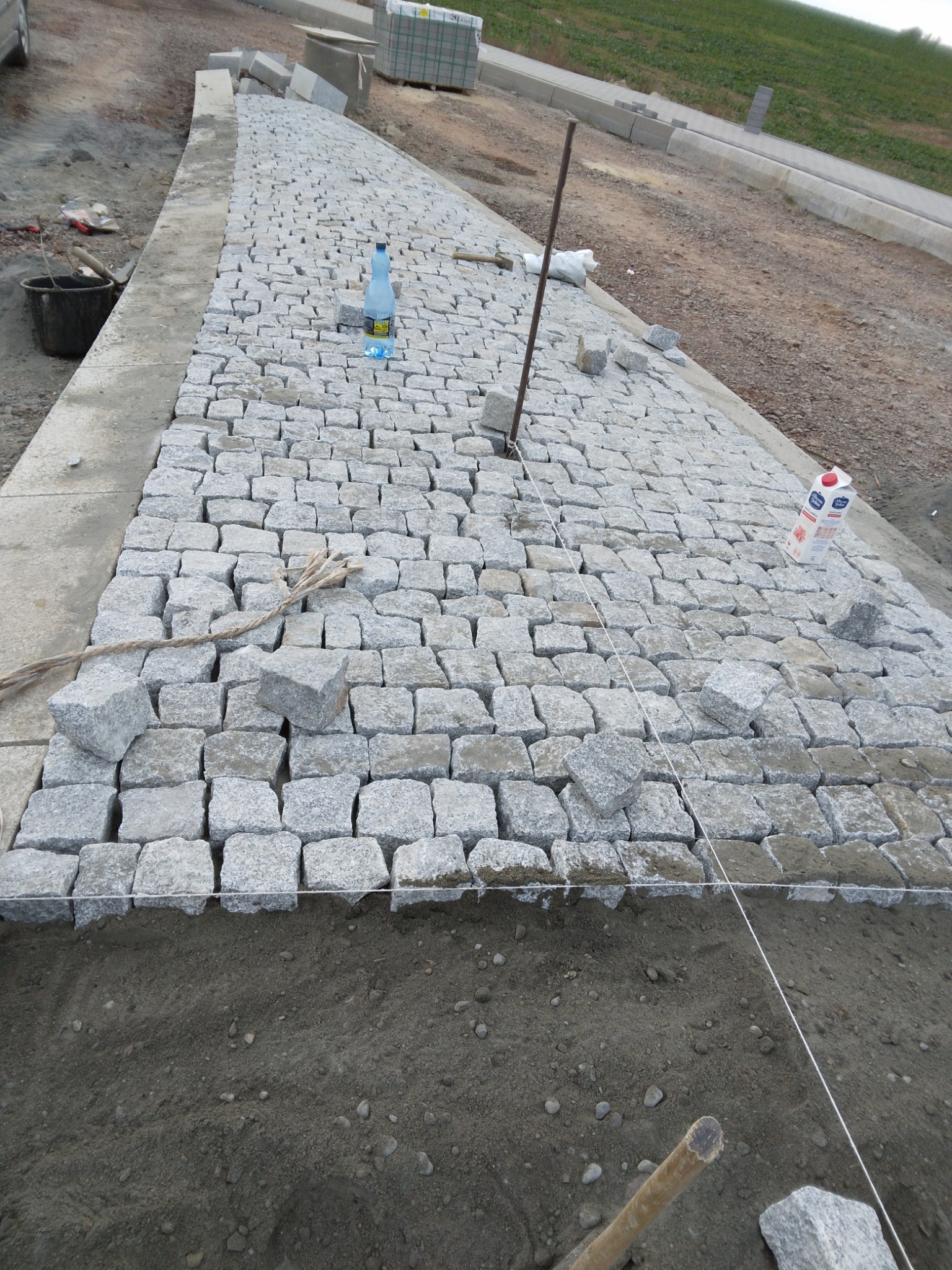 Fragment układanej ścieżki z kostki granitowej, widoczne sznurki wyznaczające linię, częściowo ułożona powierzchnia, przygotowane podłoże, w tle butelka z wodą i karton soku.