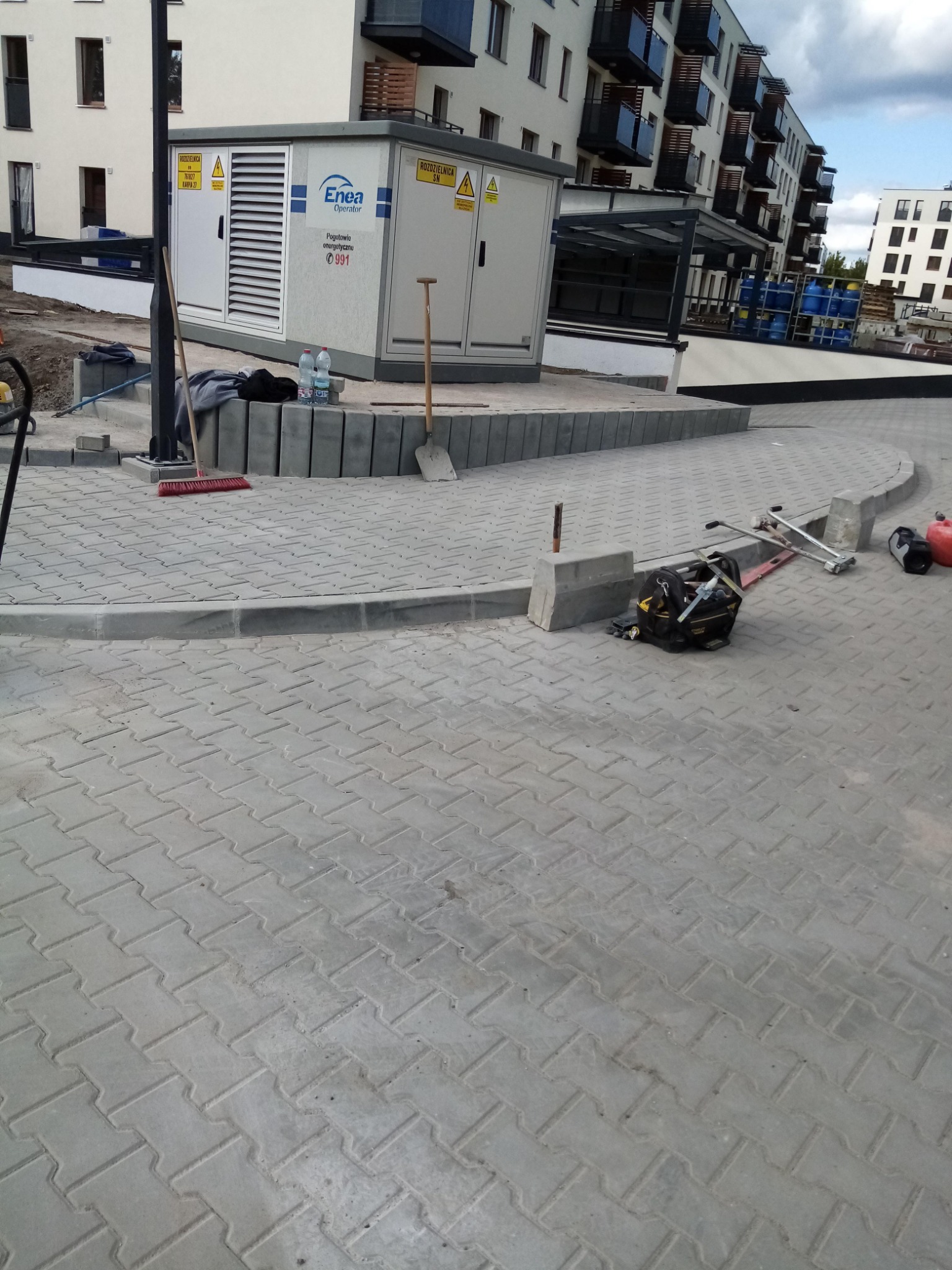 Ułożona szara kostka brukowa na placu przed budynkiem mieszkalnym, widoczne narzędzia brukarskie i elementy infrastruktury energetycznej.