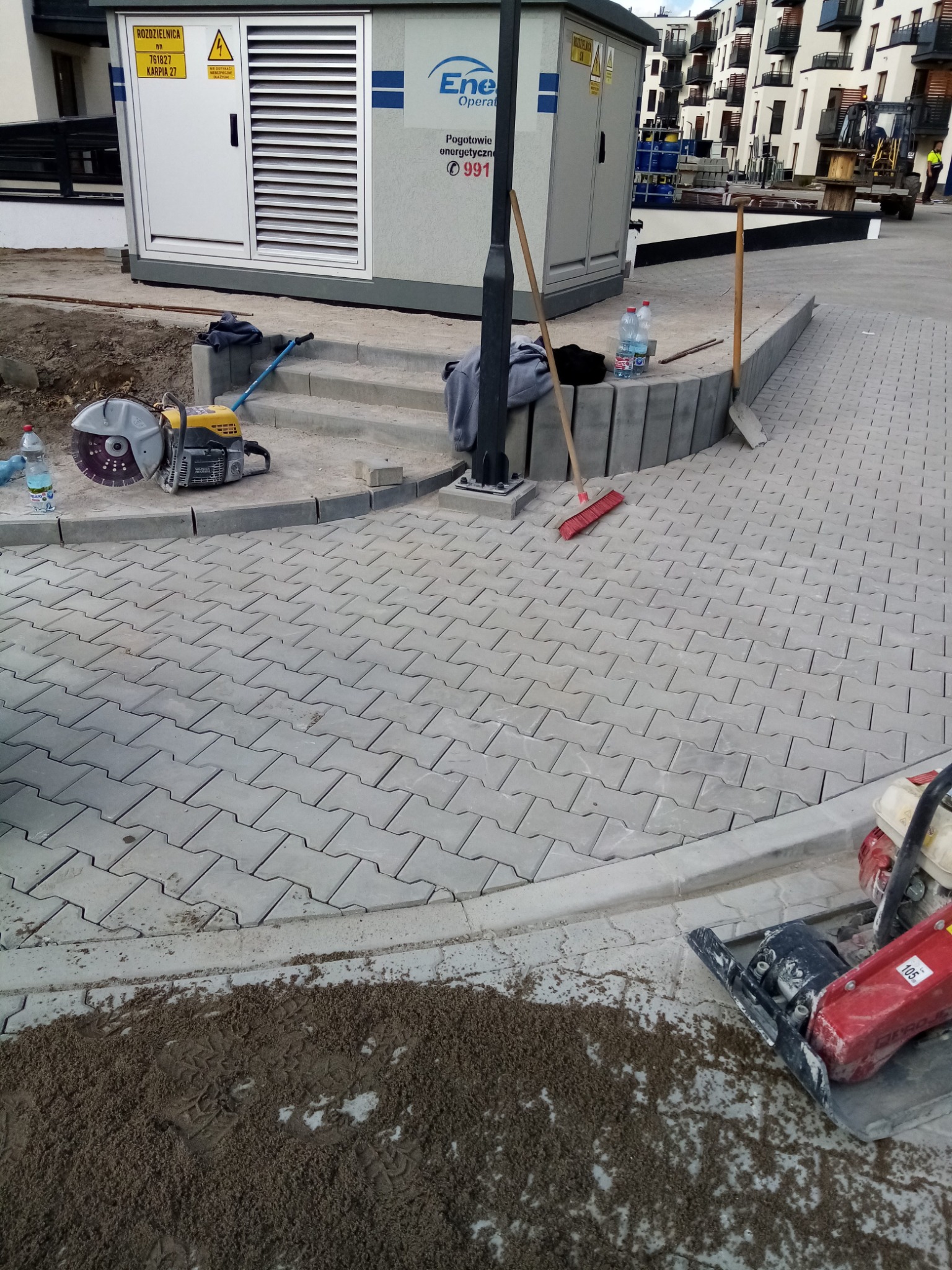 Niedokończona nawierzchnia z szarej kostki brukowej na osiedlu mieszkaniowym, widoczny sprzęt budowlany: przecinarka do kostki, zagęszczarka, szczotka i łopata, obok transformator.