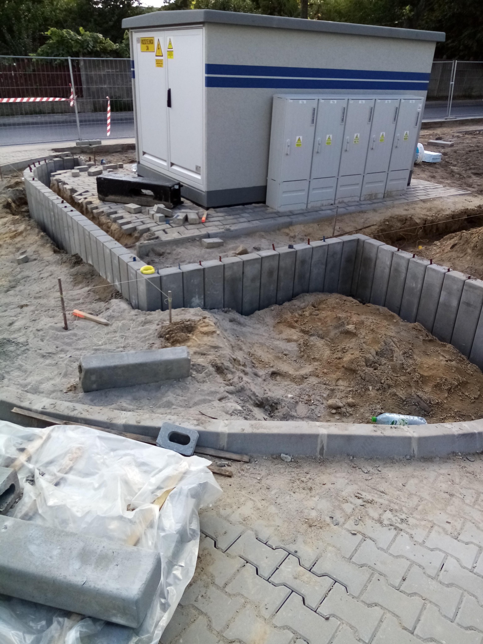 Budowa obrzeża z betonowych elementów przy transformatorze, widoczne przygotowane podłoże i częściowo ułożona kostka brukowa.