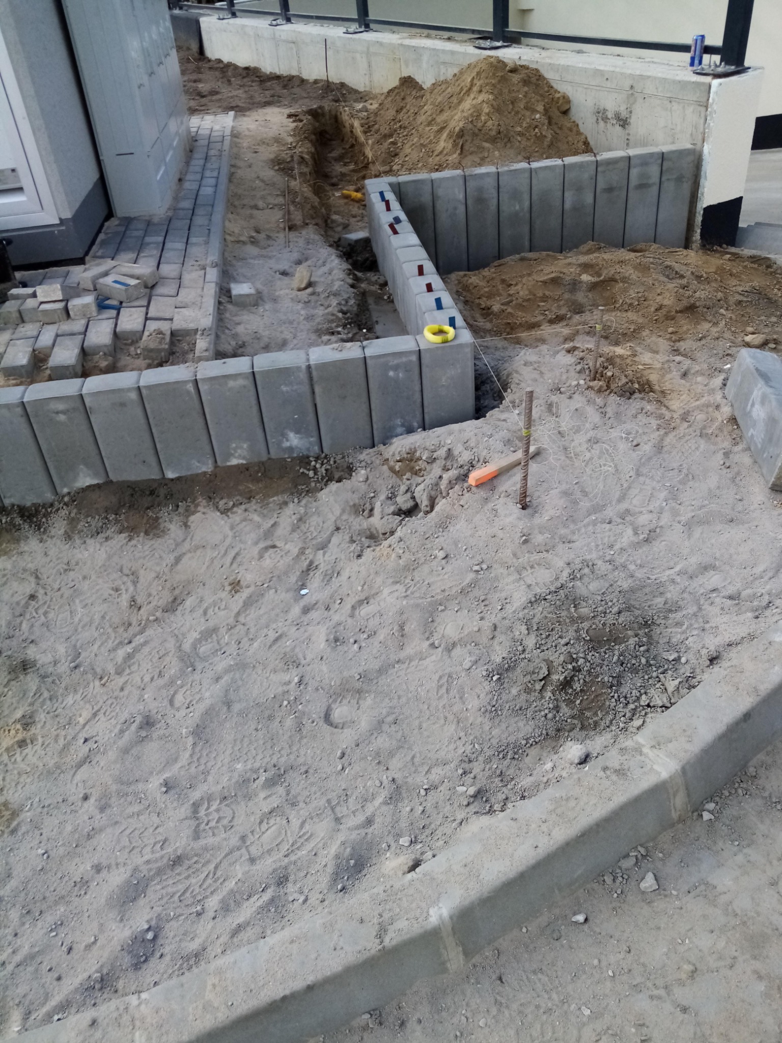 Etap prac przygotowawczych: wytyczanie terenu, krawężniki i częściowo ułożona kostka brukowa w szarym odcieniu, widoczne narzędzia i stosy piasku.