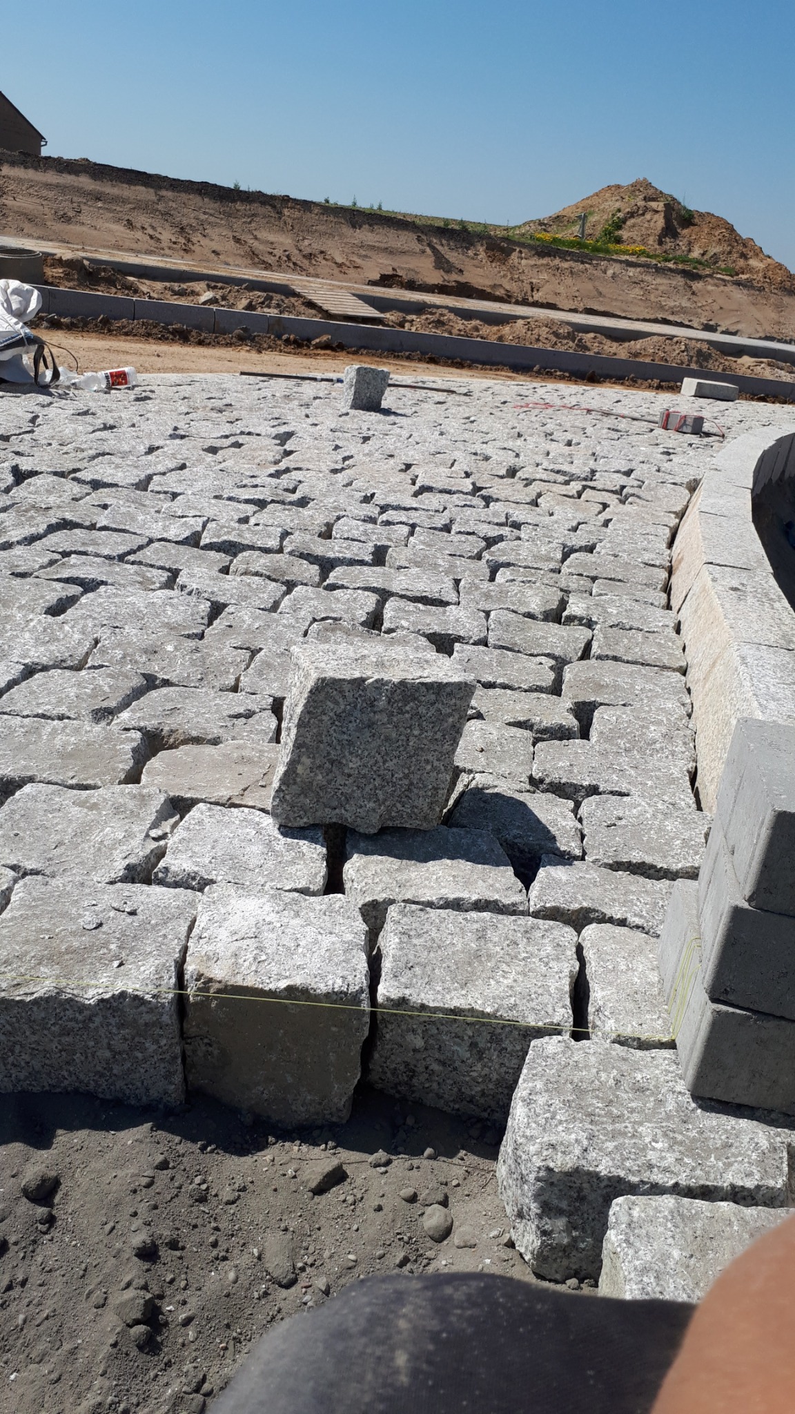 Fragment układanej nawierzchni z jasnoszarej kostki granitowej, z pojedynczym kamieniem wystającym ponad poziom, widoczne wytyczne sznurkowe i przygotowane podłoże.